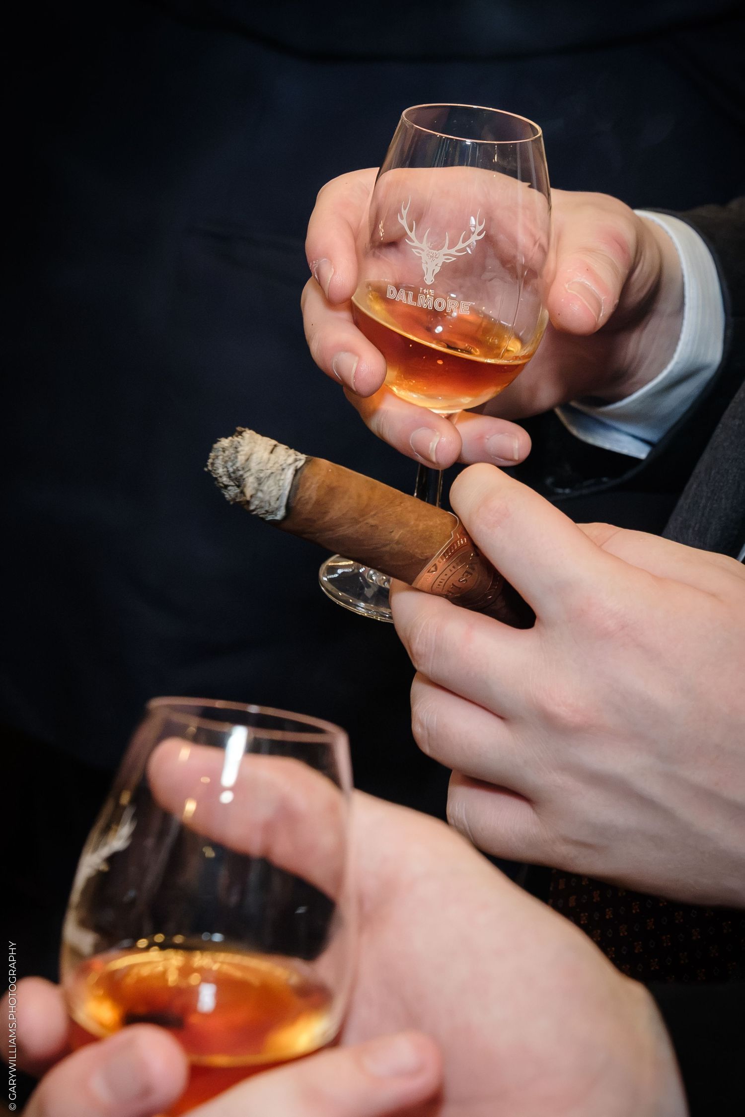 Hands holding Dalmore whisky glasses and a lit cigar at an upscale gathering.