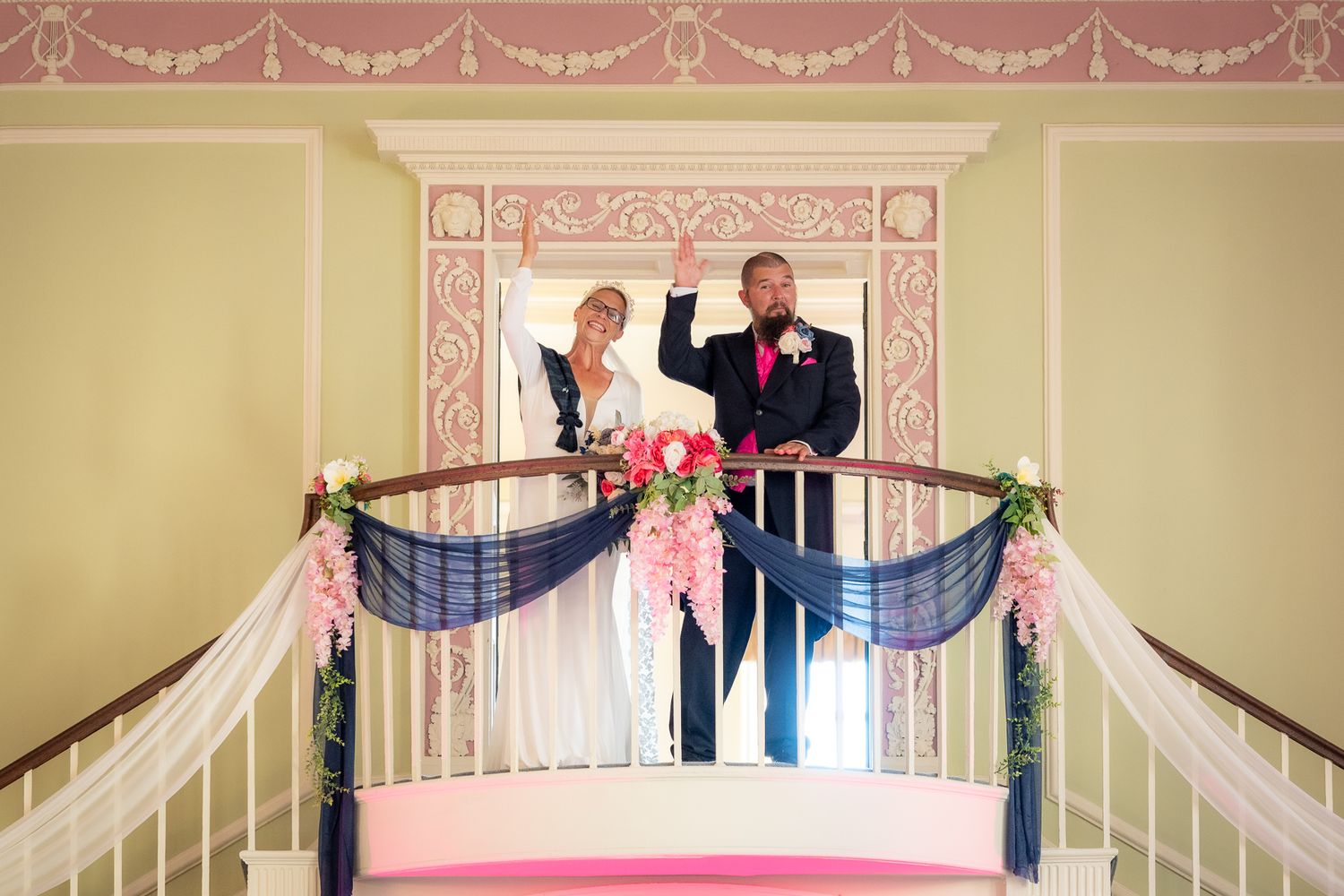 Bride and groom waving from elegant staircase balcony decorated with navy draping and pink floral arrangements.