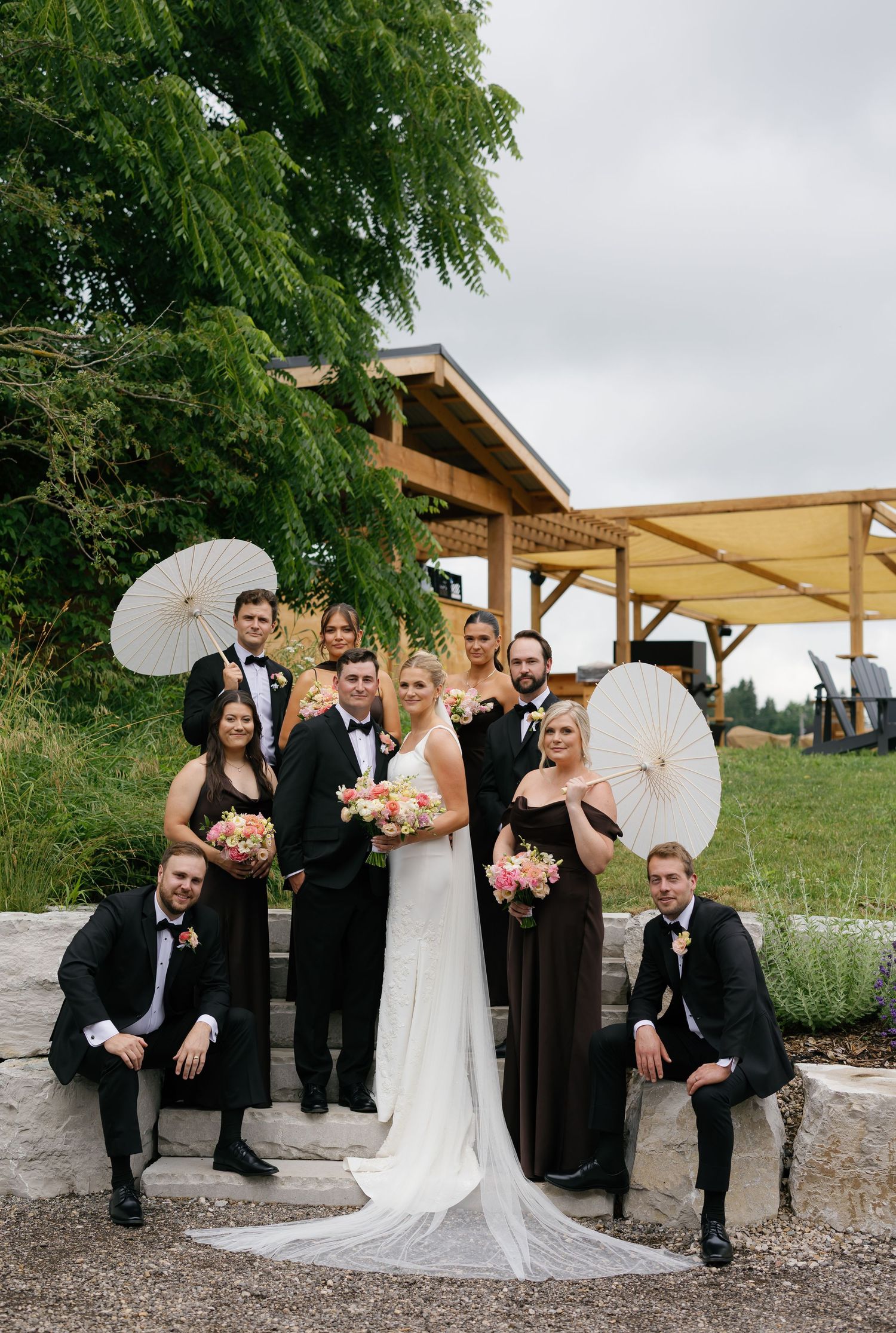 Wedding party poses outdoors with parasols, bride in white gown, bridesmaids in brown dresses, groomsmen in black tuxedos.