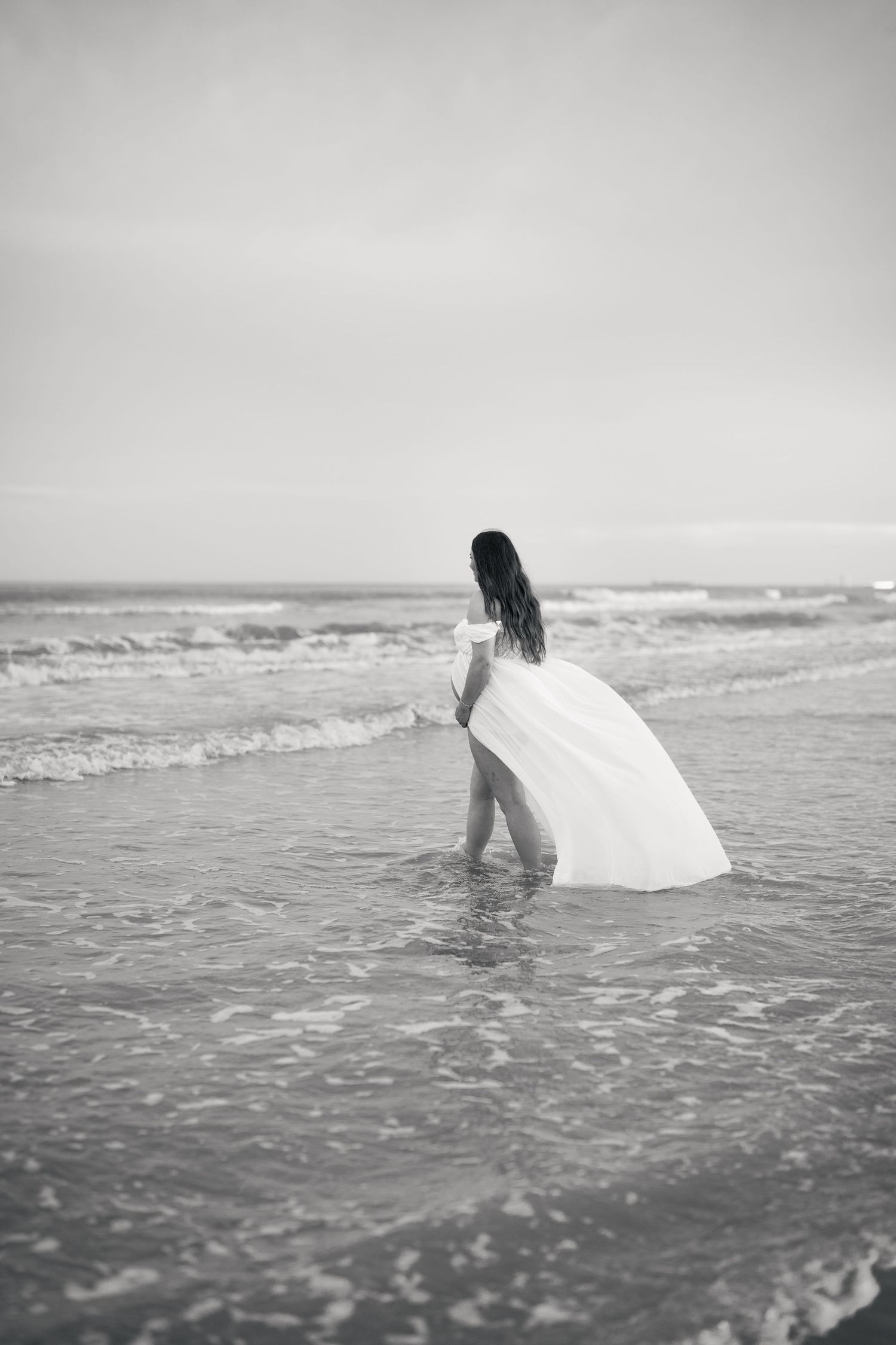Black and white photo of a woman in a flowing white dress wading into ocean waves on a beach.