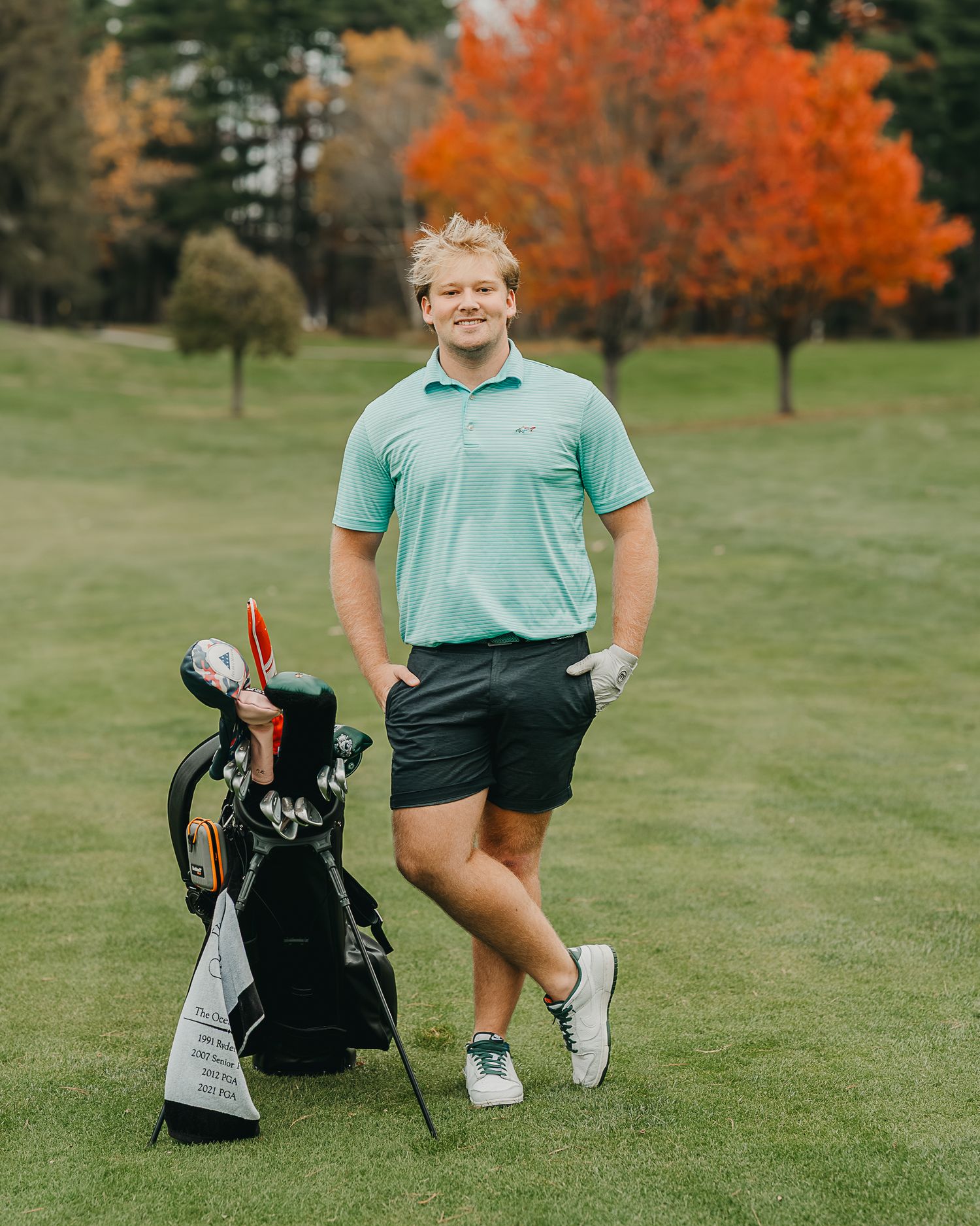 Young blonde man in mint polo shirt crouching on a golf course green, smiling confidently.