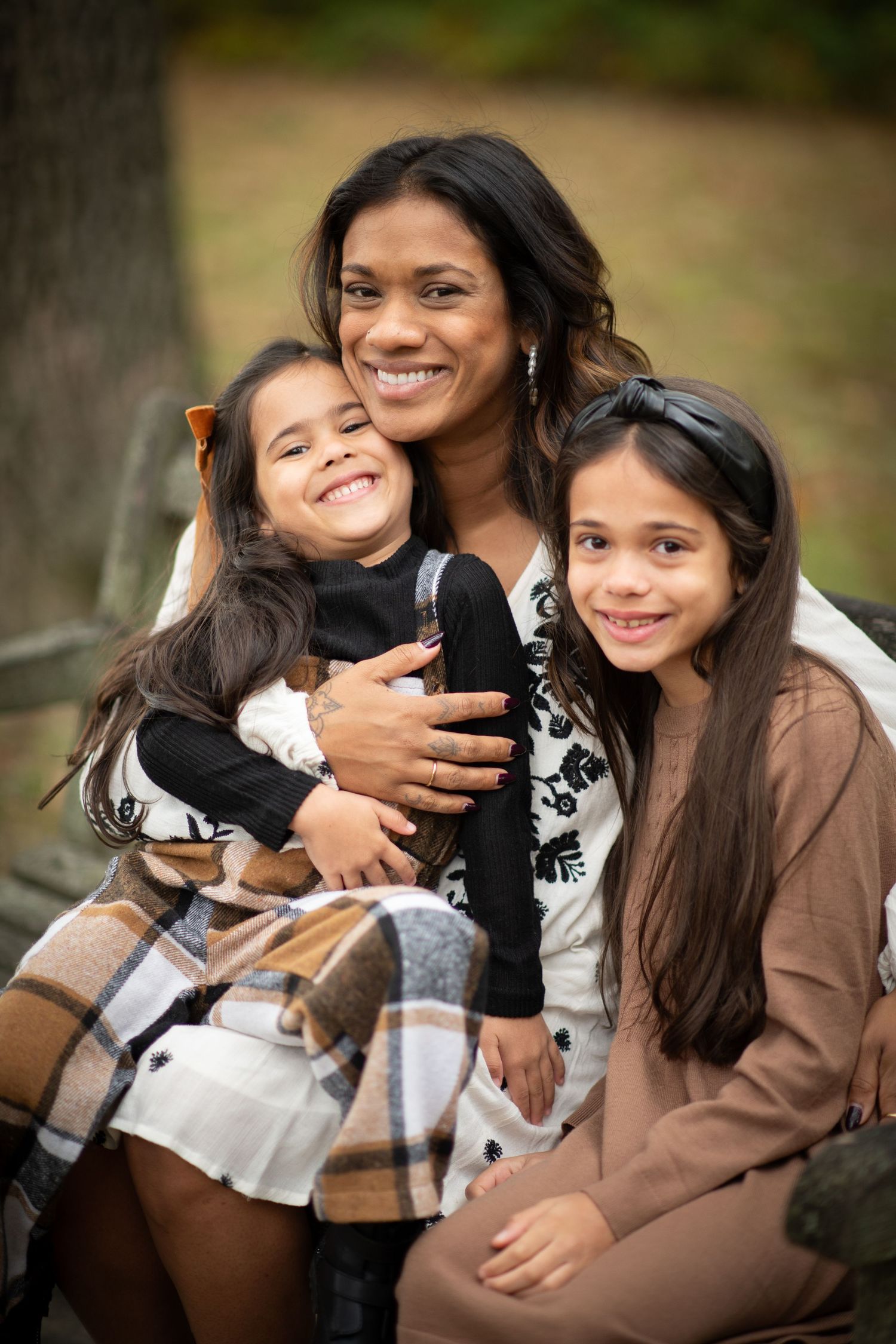 A smiling mother sits outdoors in a park with her two young daughters, sharing a warm embrace during autumn.