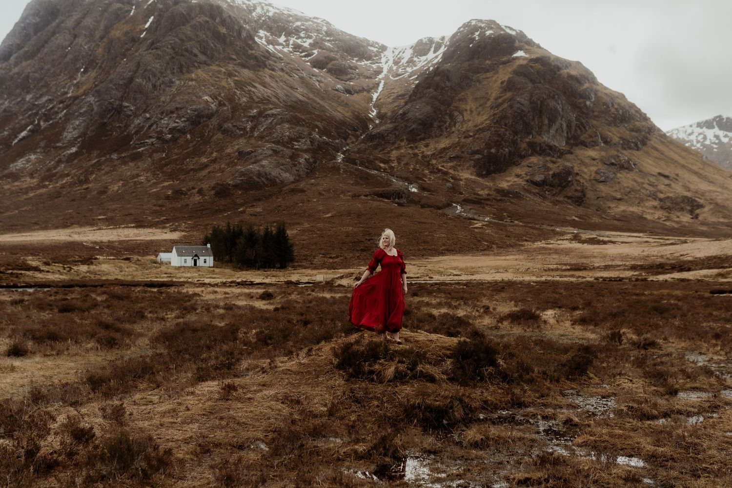 Woman in red dress stands on rocky terrain with snow-capped mountains and white cottage in misty Scottish Highlands.