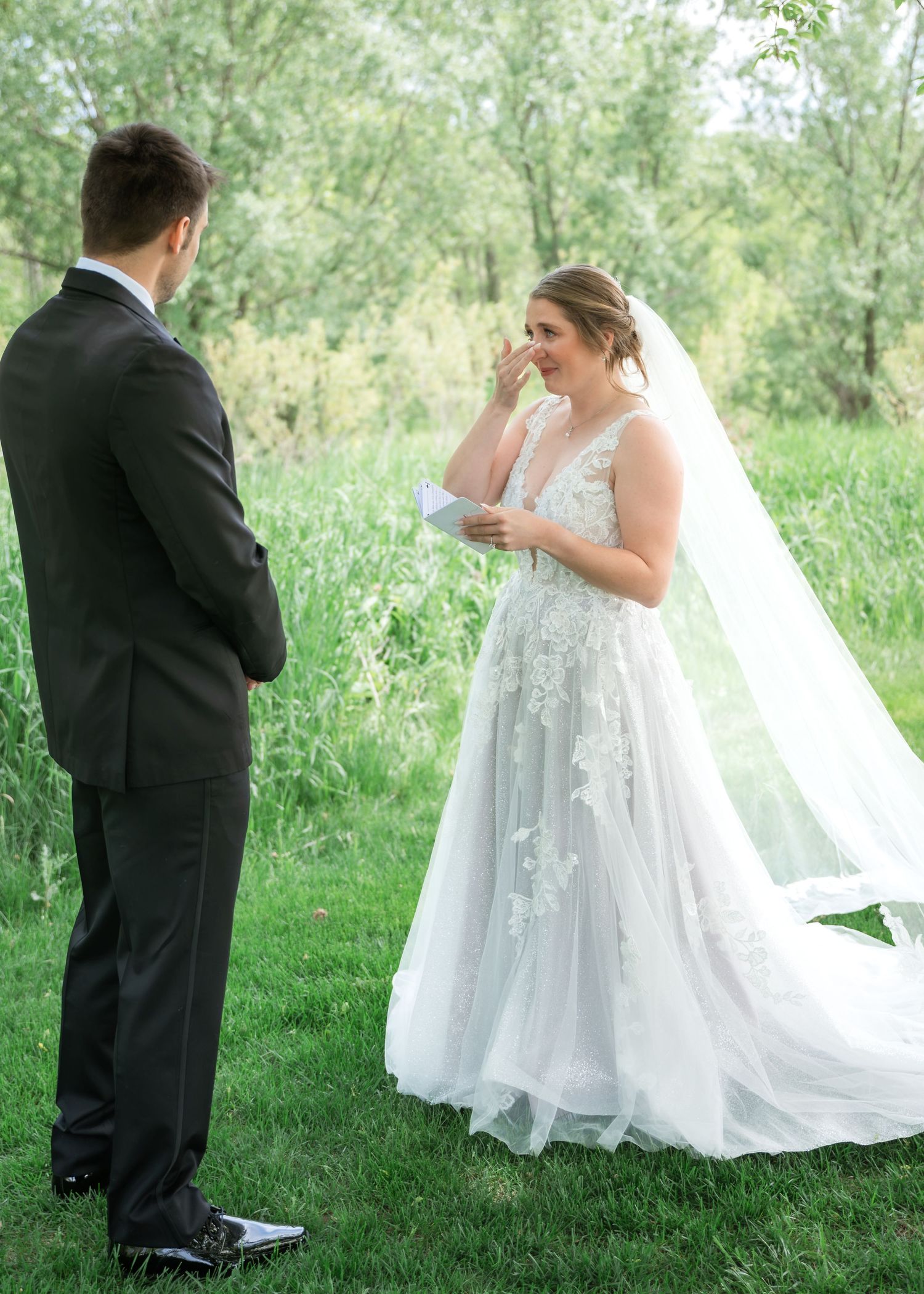 Emotional bride in lace wedding gown reads vows to groom in black tuxedo during outdoor garden ceremony.