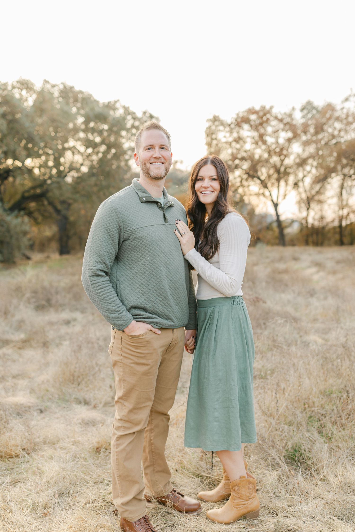 A couple poses together in a golden field, wearing coordinated earth tones and sage green, smiling warmly at the camera.