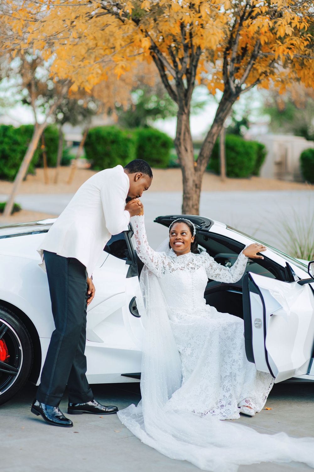 Groom in white suit kisses bride's hand as she exits a white luxury convertible in a lace wedding gown.
