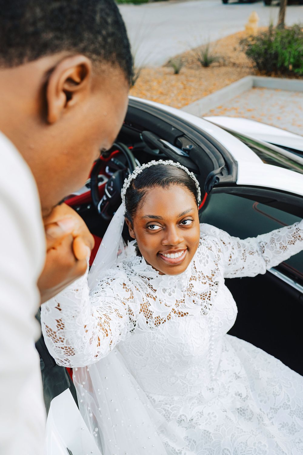 A smiling Black bride in white lace gown and veil exits a car, holding hands with groom in white suit.