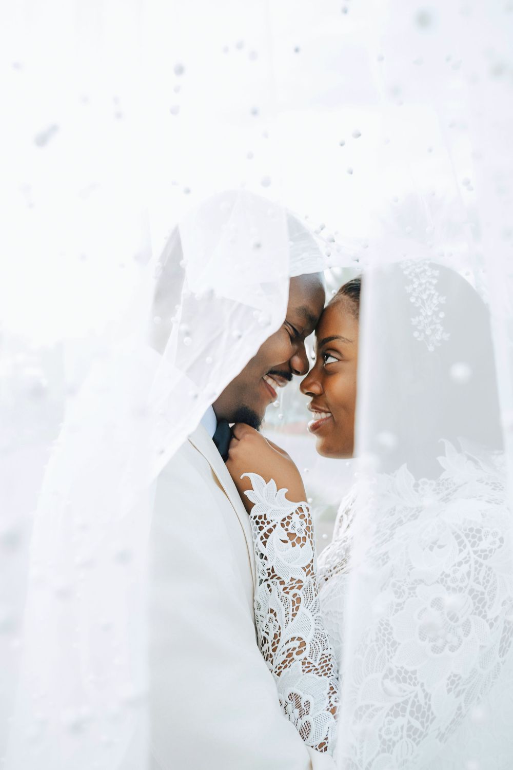 Smiling Black newlyweds share intimate moment under white dotted veil in bright wedding portrait.