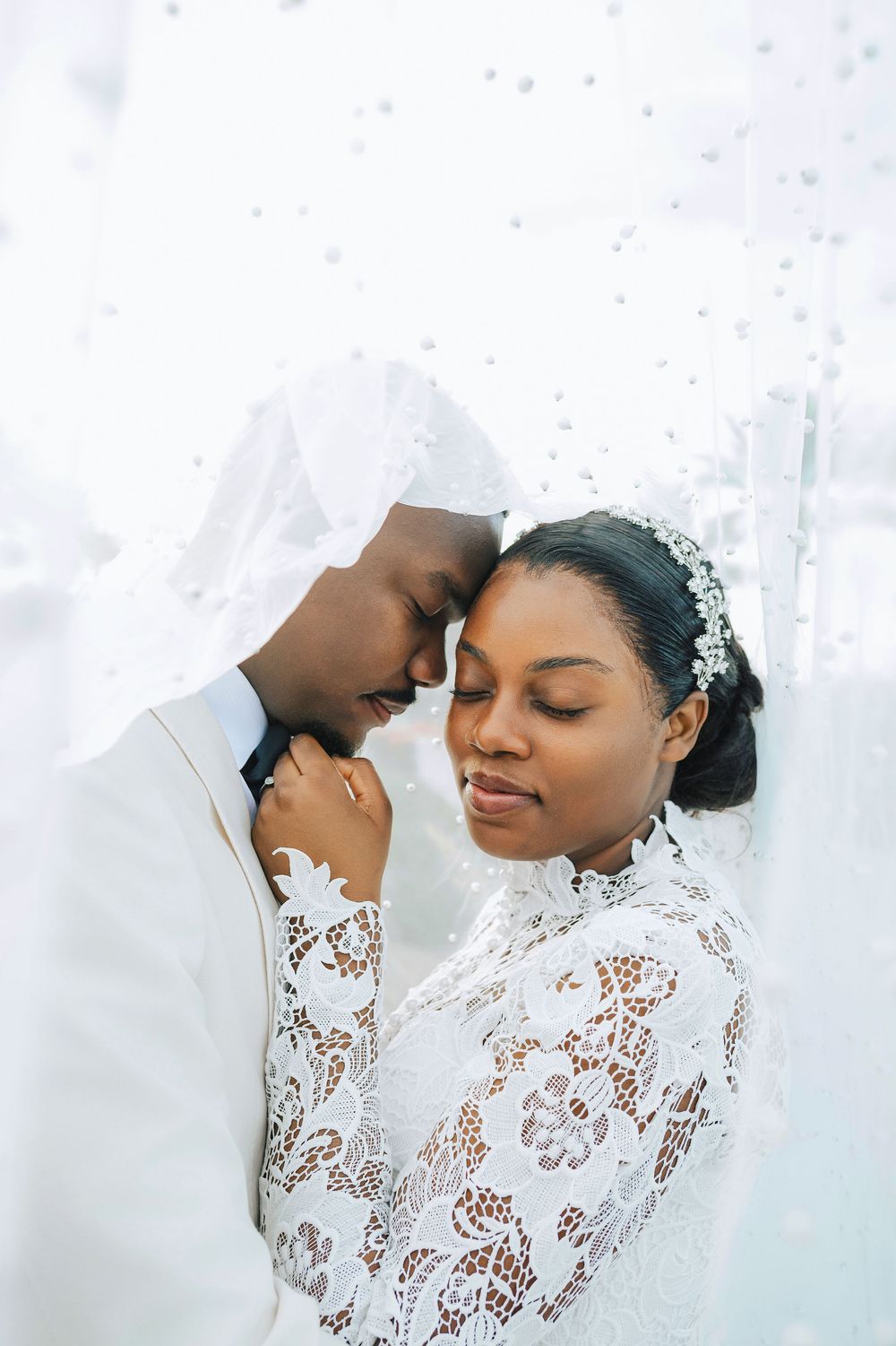 Black bride and groom touch foreheads tenderly under pearl-dotted white veil in romantic portrait.