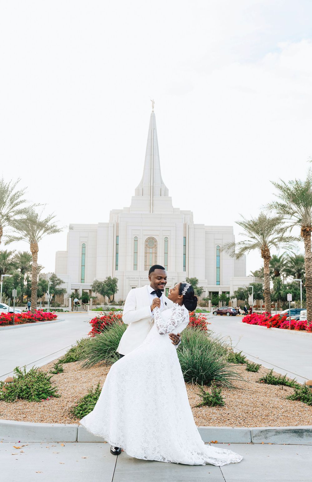 Black bride and groom embrace in front of white LDS temple with red flowers and palm trees.