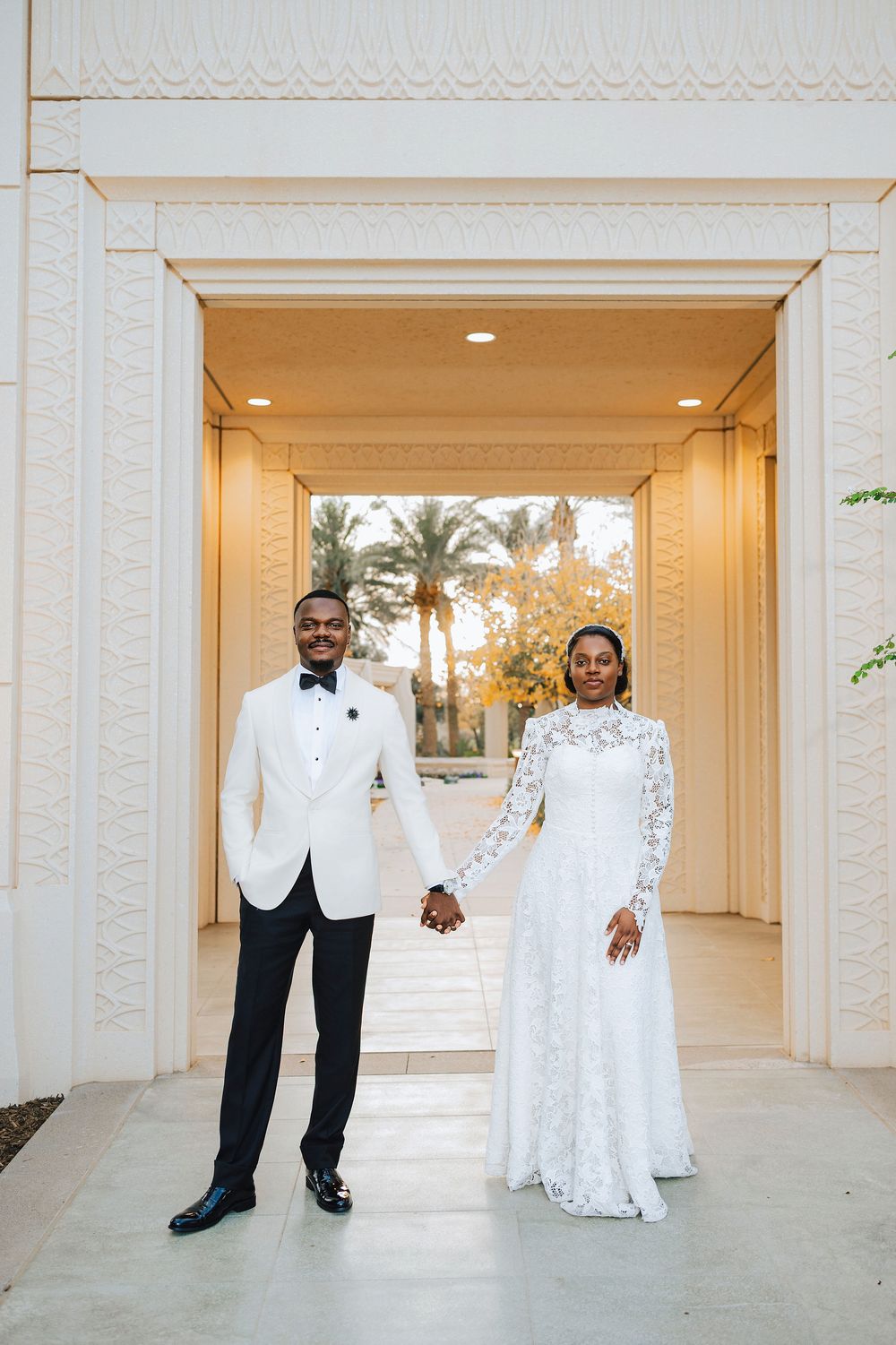 A elegant Black couple holds hands in wedding attire at a beautiful archway entrance.