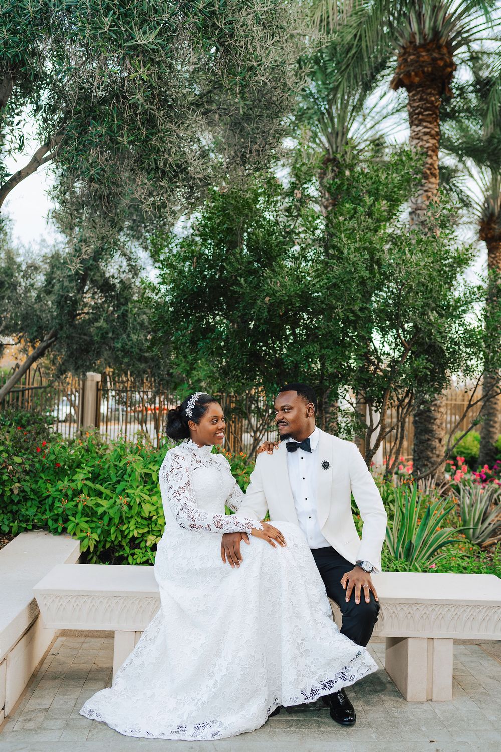 Black couple in white wedding attire sit on bench surrounded by lush greenery and palm trees.