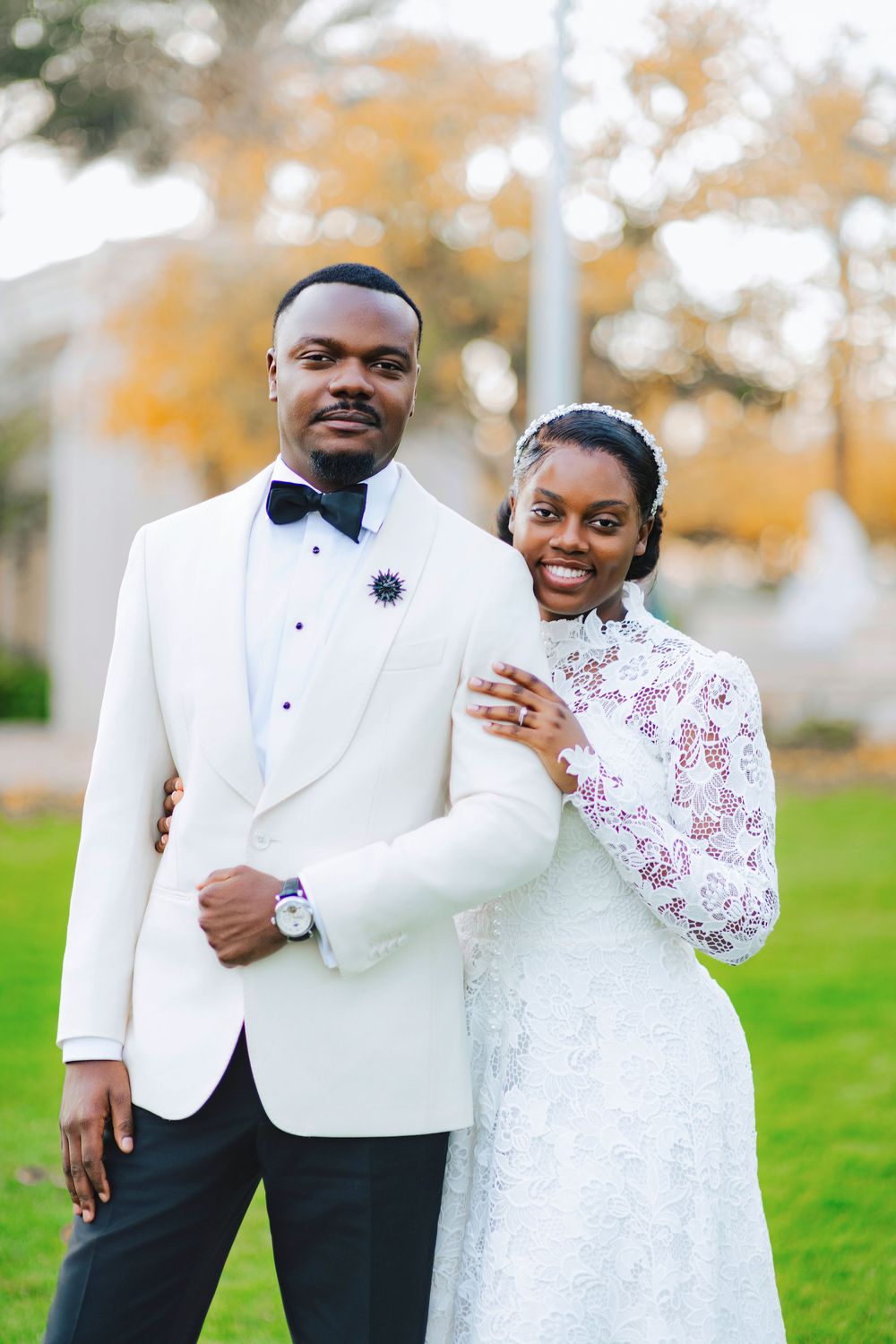 Black couple in wedding attire outdoors, groom in white tuxedo, bride in lace high-neck gown, autumn trees behind.