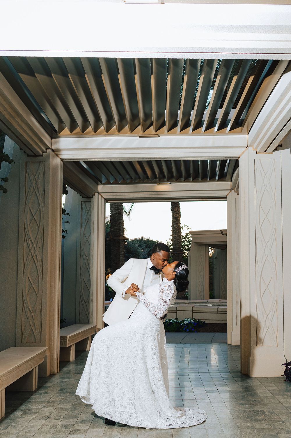 Bride and groom share a romantic dip kiss under a pergola in matching white wedding attire.