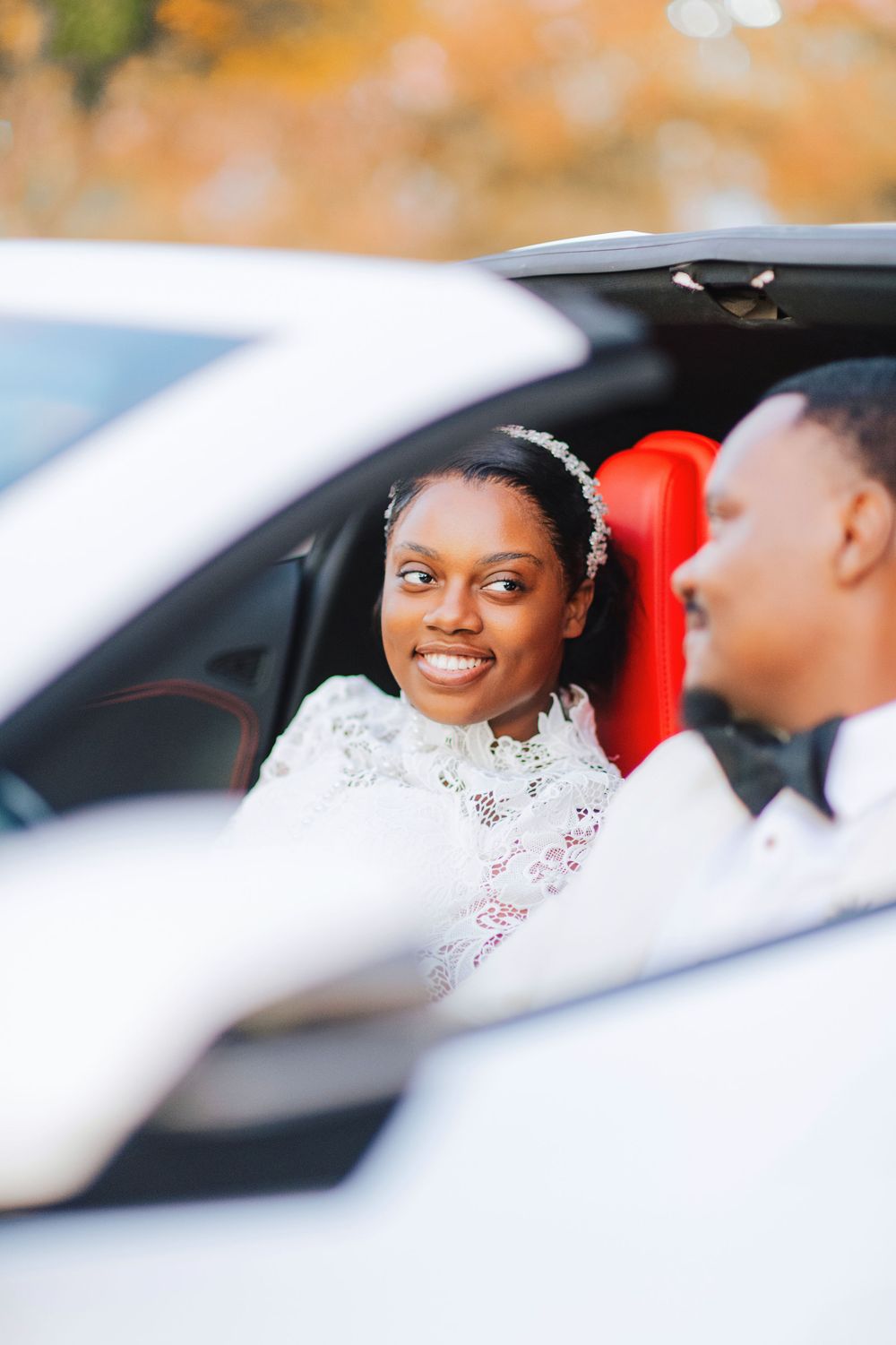 Smiling Black bride in lace gown sits in white luxury car with groom during wedding day photoshoot.