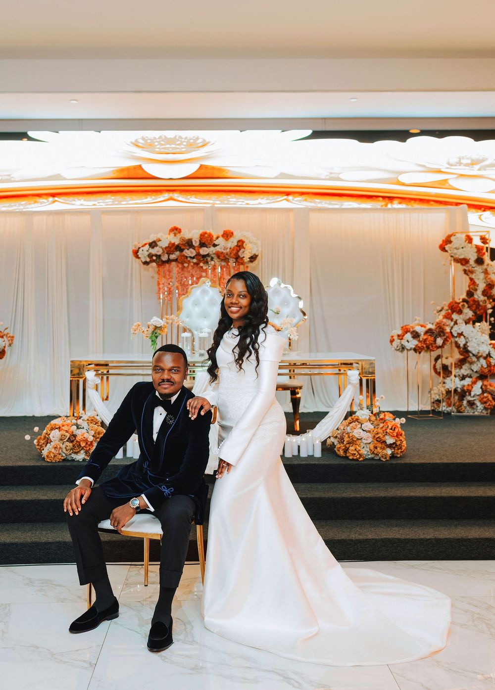 Elegant Black couple poses at wedding reception, groom in tuxedo, bride in white long-sleeve gown, gold floral backdrop.