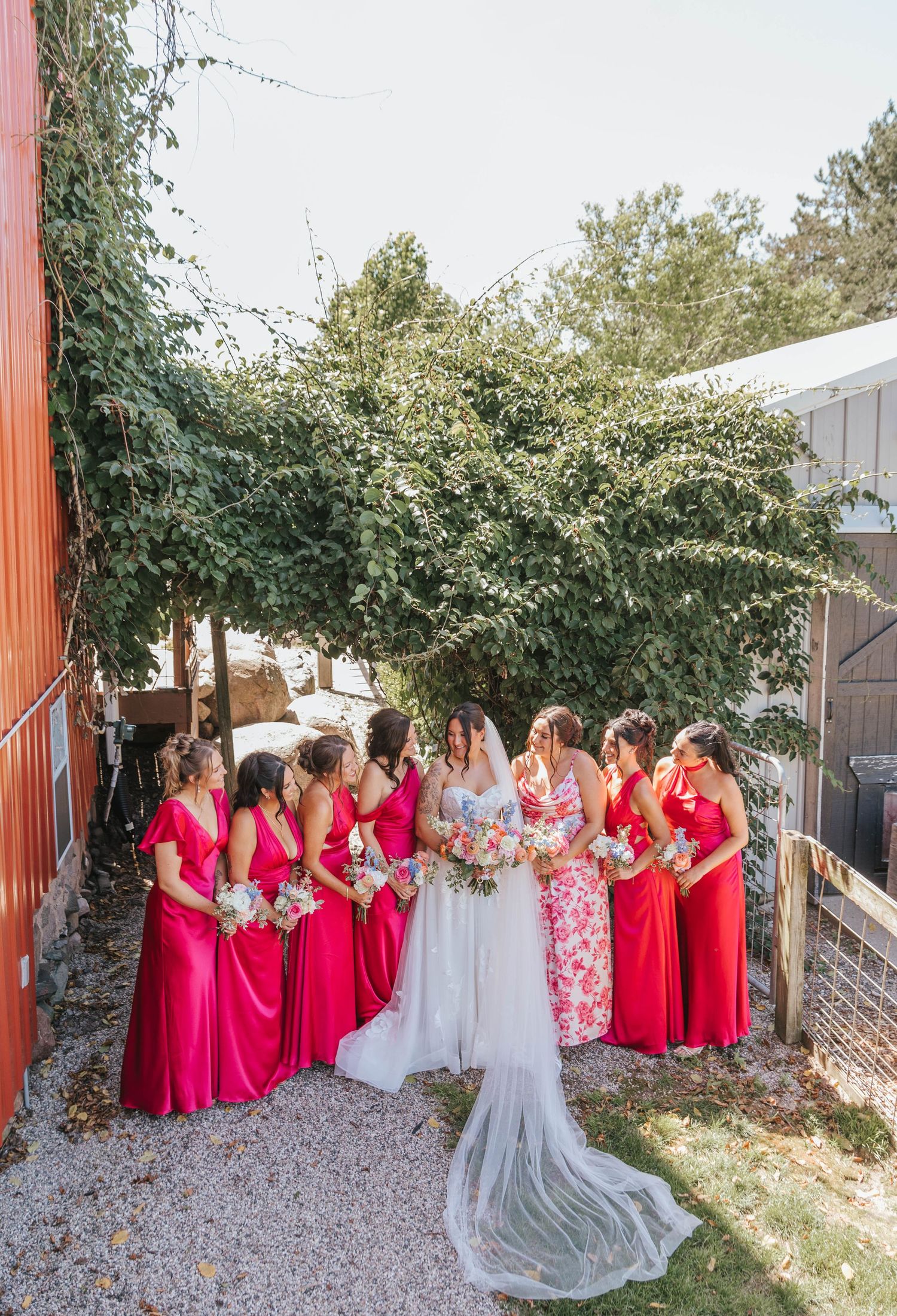 Bride in floral gown with bridesmaids in red dresses holding bouquets outdoors near ivy-covered wall.