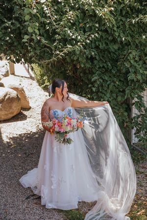 Bride in floral strapless wedding gown holding colorful bouquet while flowing veil billows beside lush green trees.