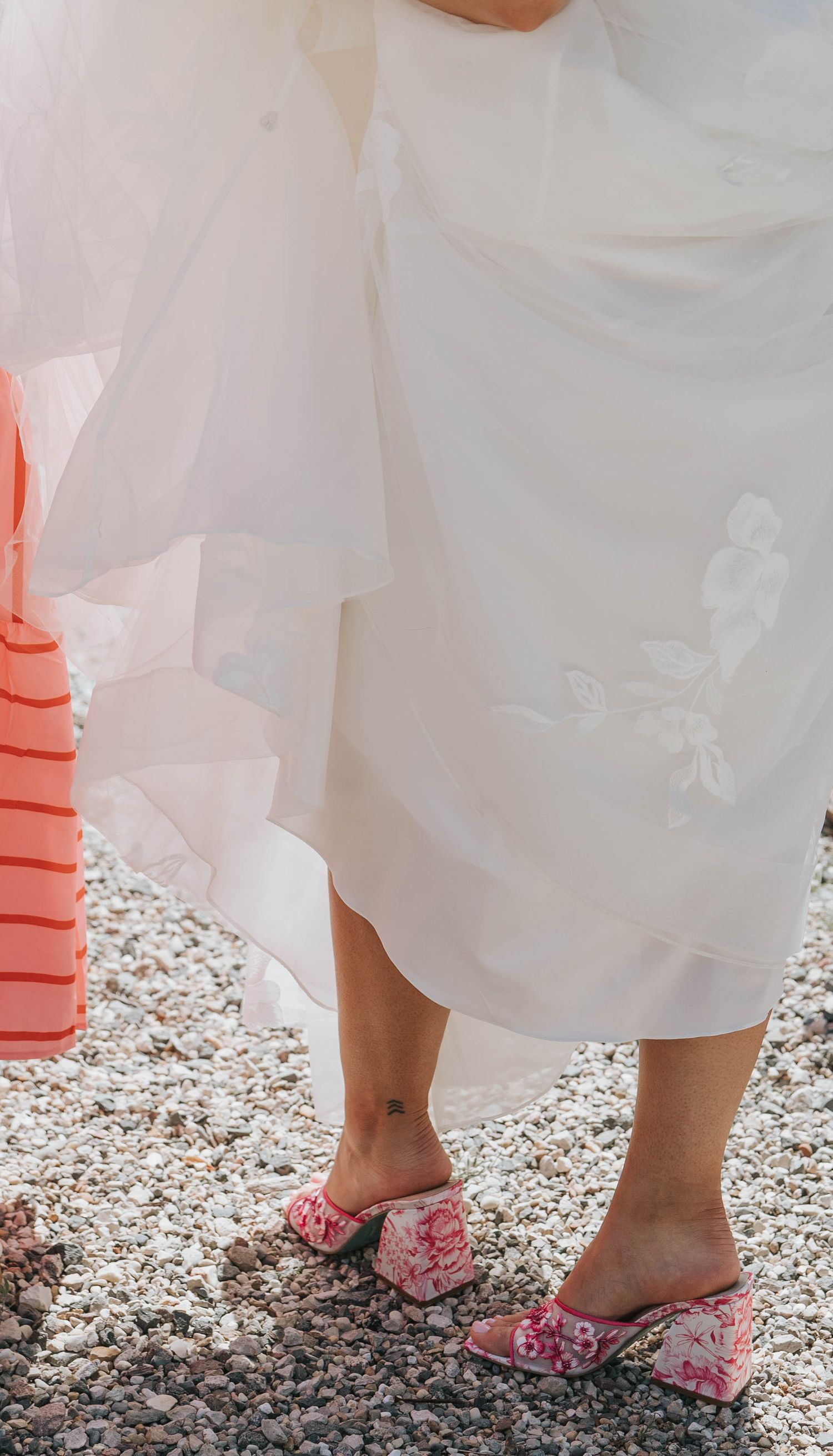 Bride in white wedding dress wearing pink floral mule heels standing on gravel, with pink striped bag visible.