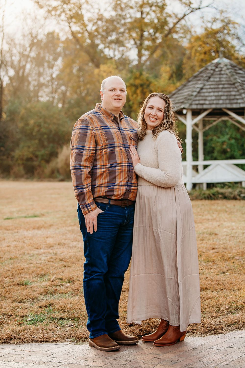 Smiling couple embracing near a white gazebo on a brick path in an autumn park.