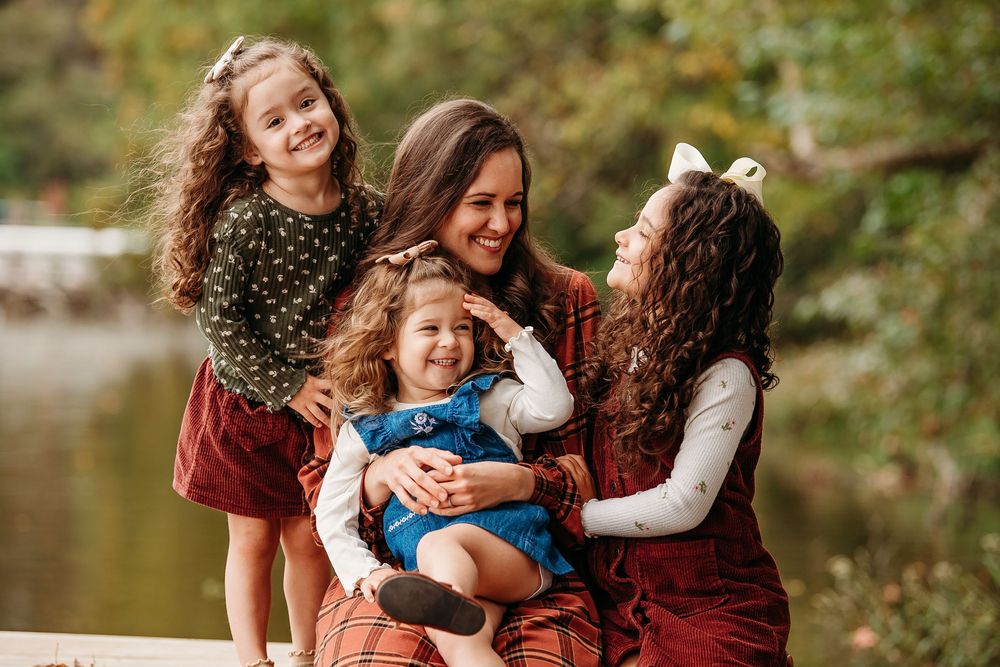Mother smiling with three curly-haired daughters on a lakeside dock in autumn.