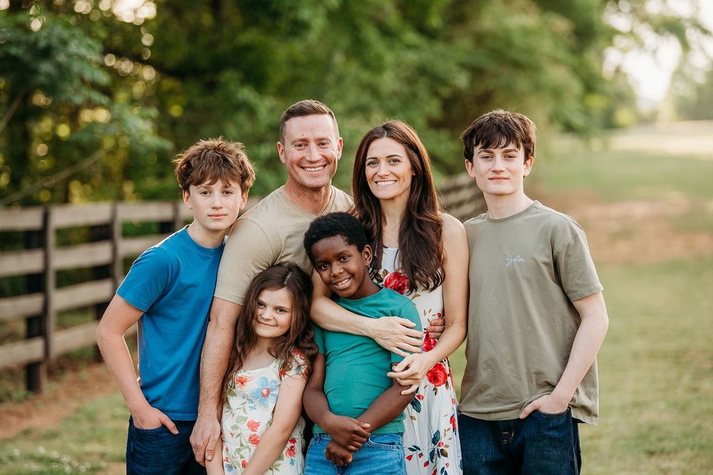 Smiling family of six posing together in a sunny green field near a wooden fence.