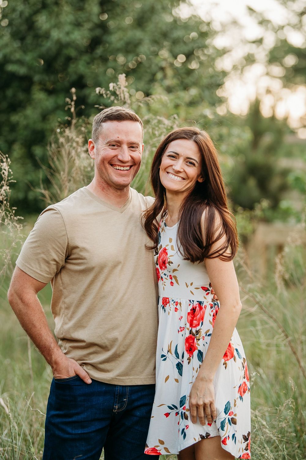 Smiling couple posing outdoors in a green field during golden hour, man in tan shirt and woman in floral dress.