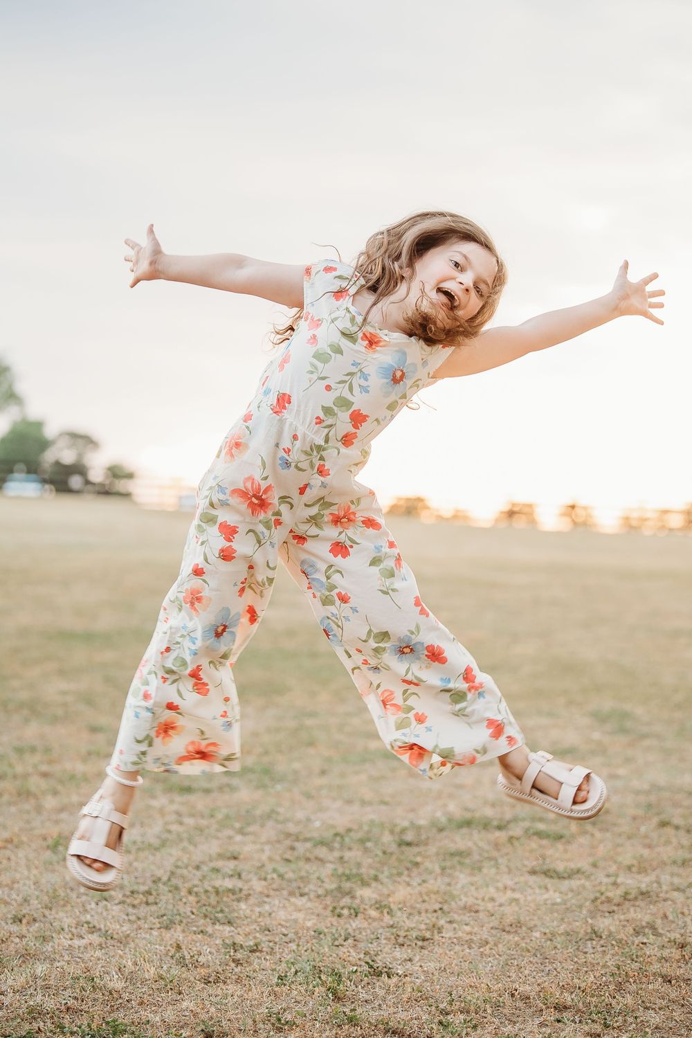 Joyful young girl in a floral jumpsuit jumping with arms outstretched in an open field at sunset.