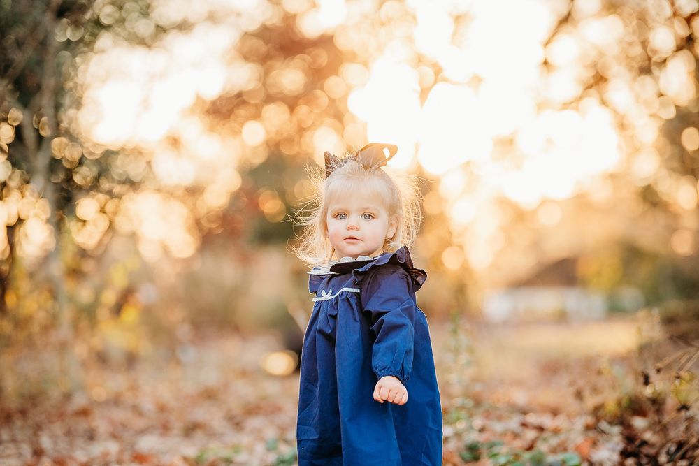 Cute blonde toddler girl in a navy blue coat standing among fall foliage in warm golden light.