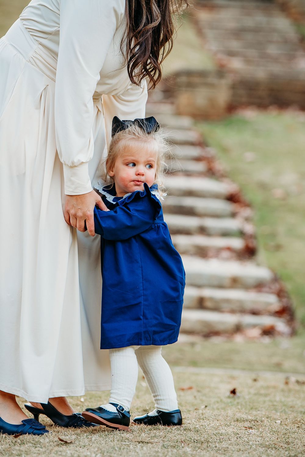 Adorable toddler girl in a blue dress clinging to her mothers white gown near stone garden steps.