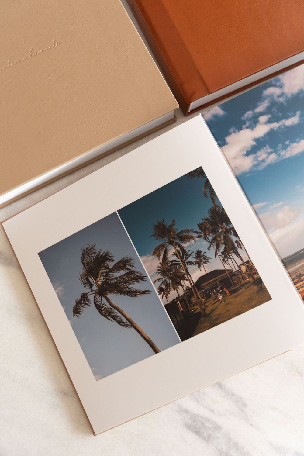 Photo album open to a page with two tropical palm tree photos on white matting, beside a brown leather album box.