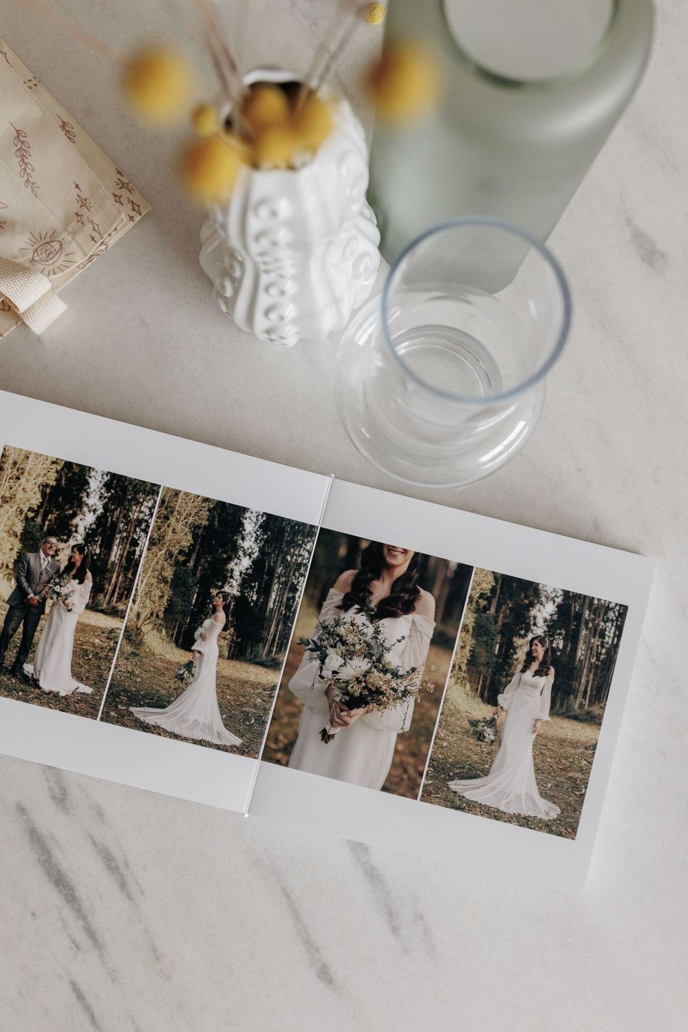 Open wedding photo album displaying bride portraits on marble table with vase and glass.