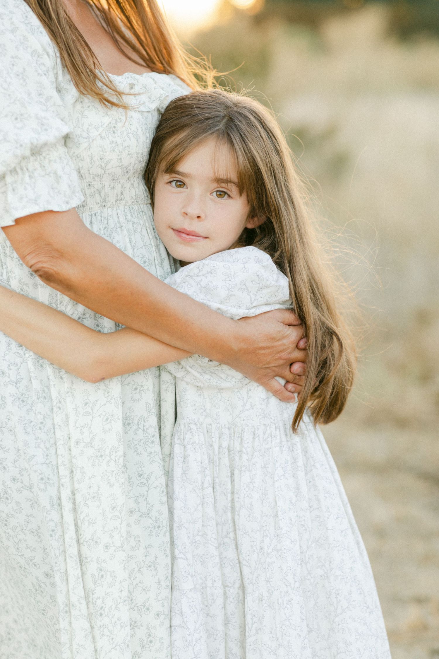 A young girl in a white floral dress hugs her mother outdoors in warm golden light.