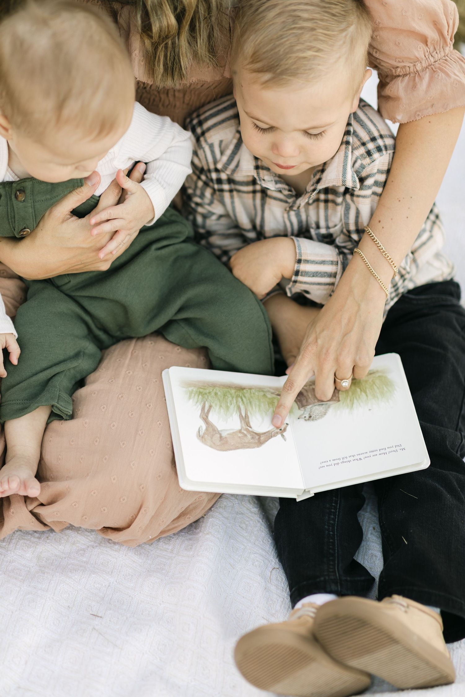 Adorable baby in green overalls lying in a wicker basket, looking up curiously while an adult gently touches their cheek.