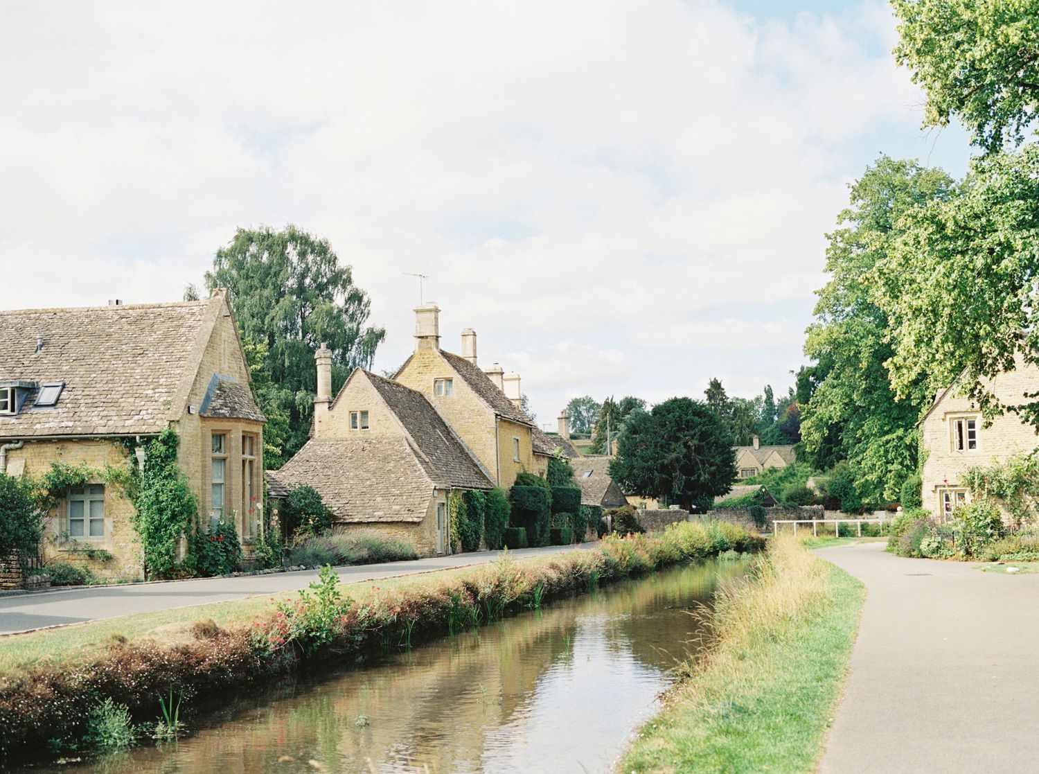 Charming Cotswolds village with stone cottages lining a calm stream, surrounded by lush green trees under a cloudy sky.
