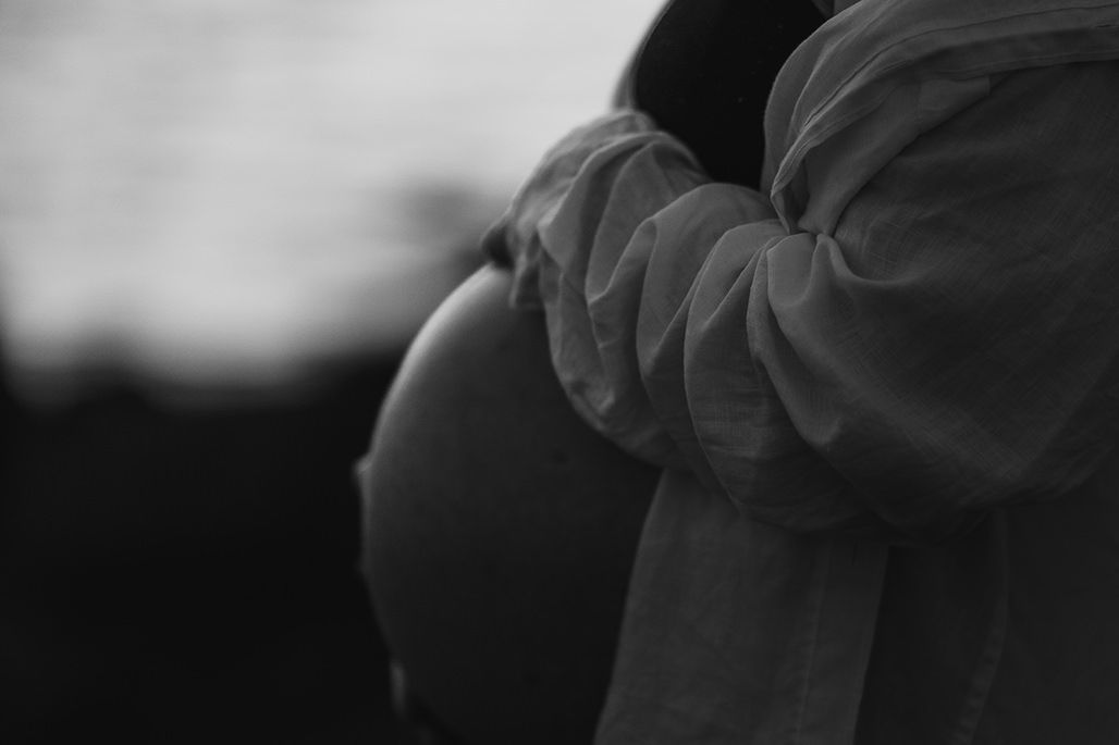 Black and white photo of a pregnant woman being embraced by her partner near the ocean shore.