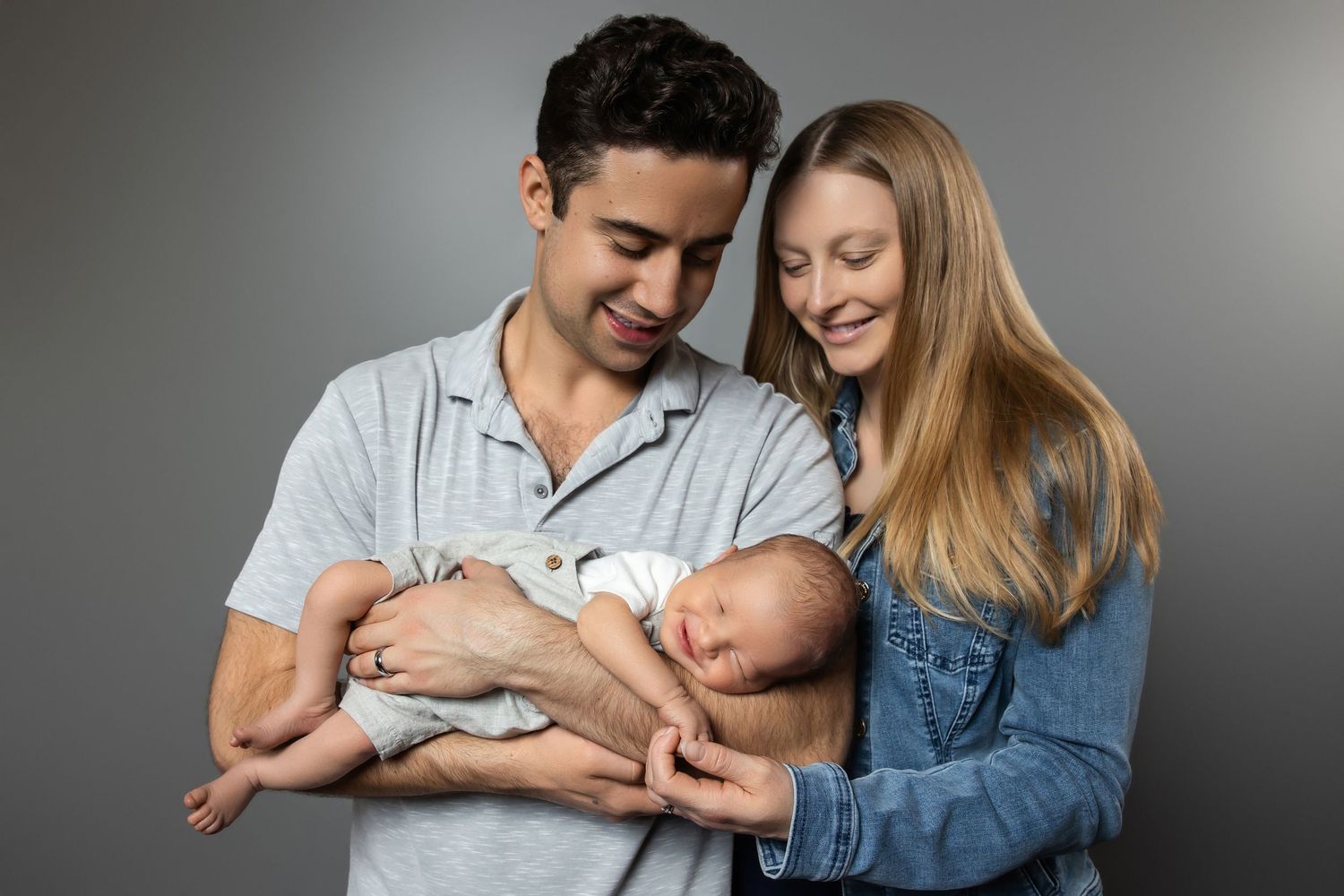 A happy couple smiling and cradling their newborn baby against a gray studio background.