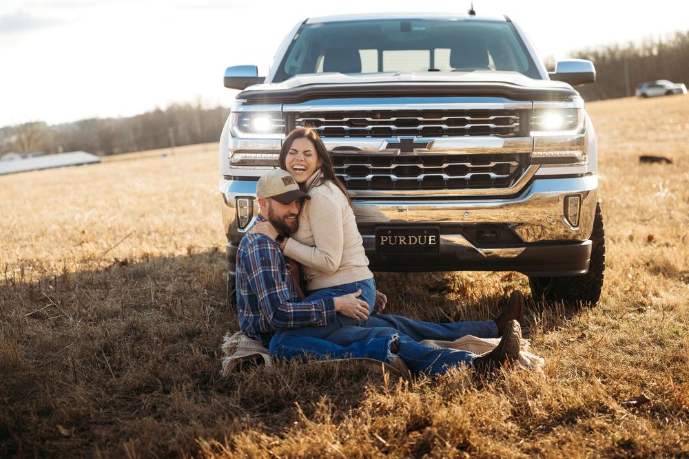 Couple sitting together in a golden field in front of a white Chevrolet truck at sunset.