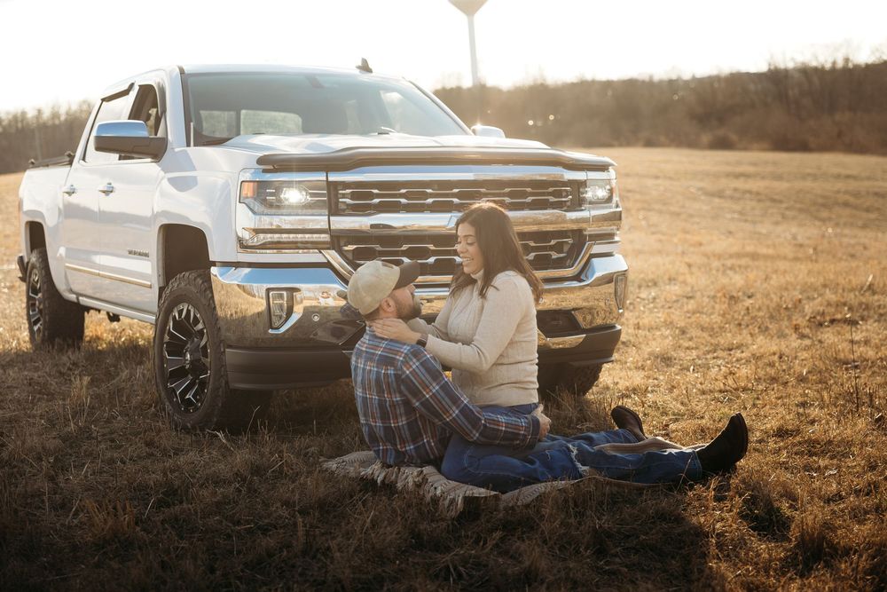 Couple sitting together in a golden field leaning against a white Chevrolet Silverado truck at sunset.