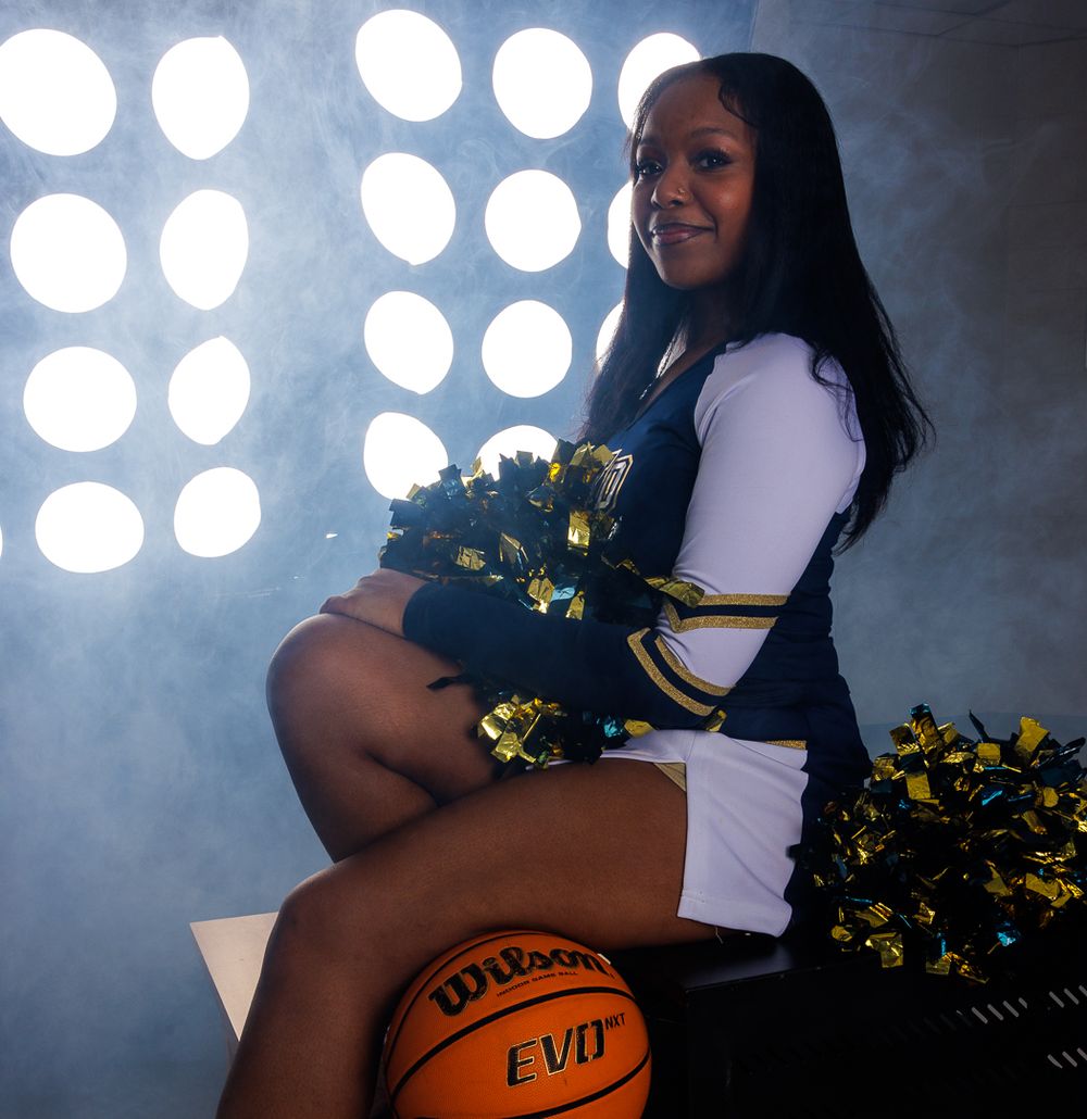 A cheerleader poses with pom poms and basketball in a dramatic studio lighting setup.