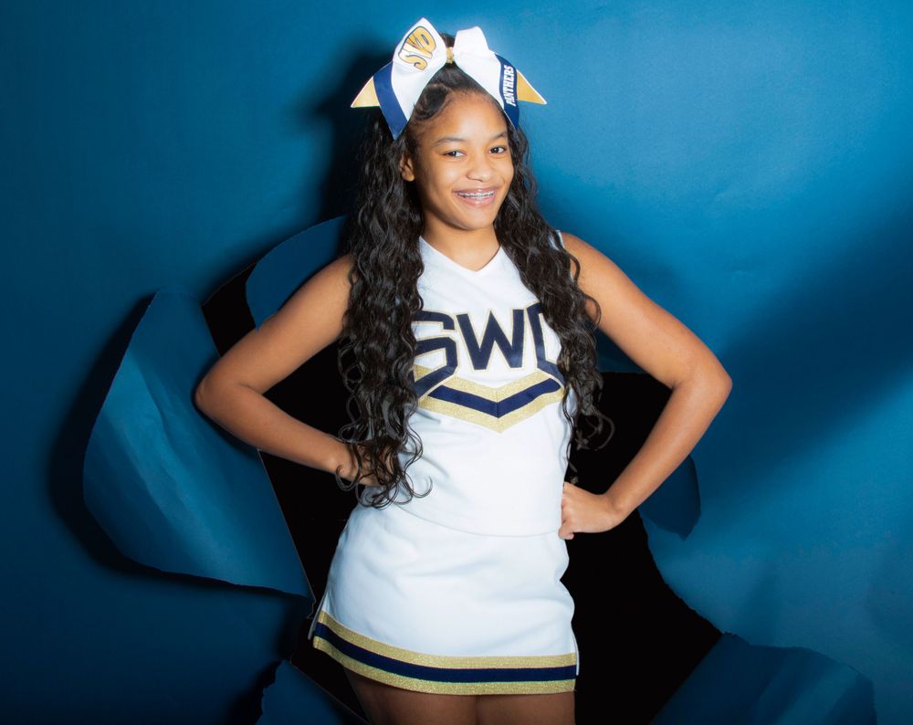 Cheerleader in white uniform with bow poses against blue backdrop.