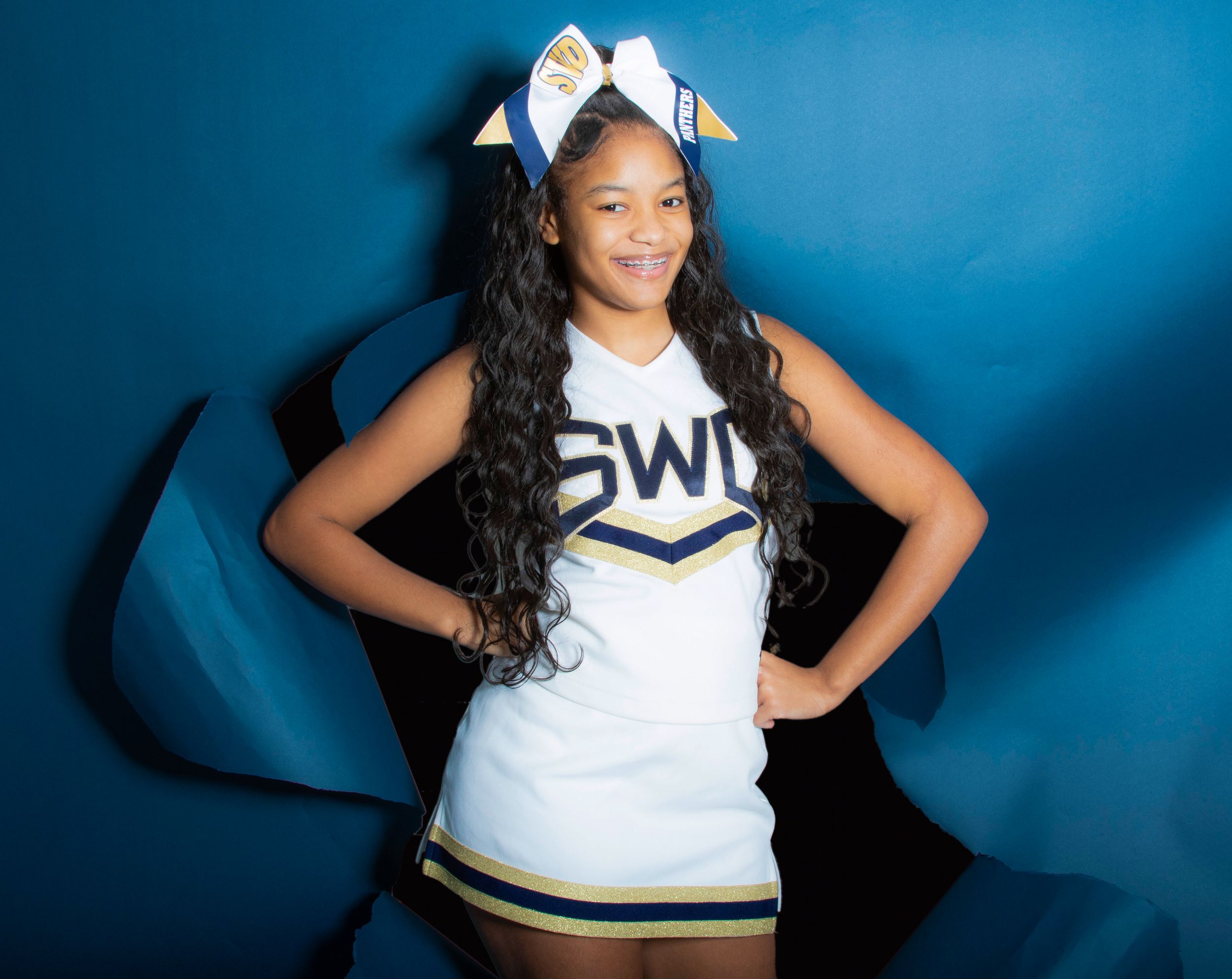 Cheerleader in white uniform with bow poses against blue backdrop.