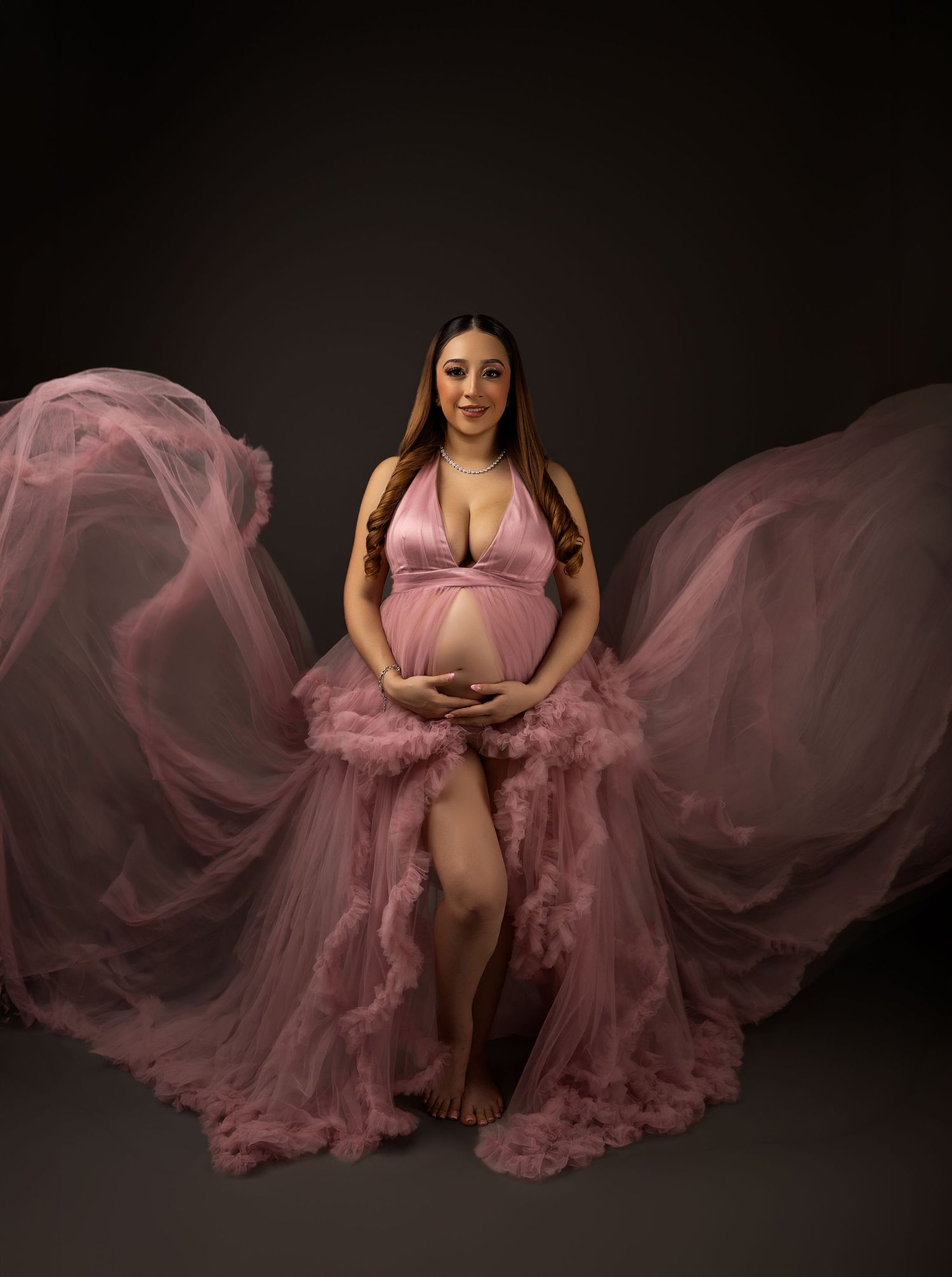 Pregnant woman in flowing pink tulle maternity gown poses gracefully in professional studio photography session.
