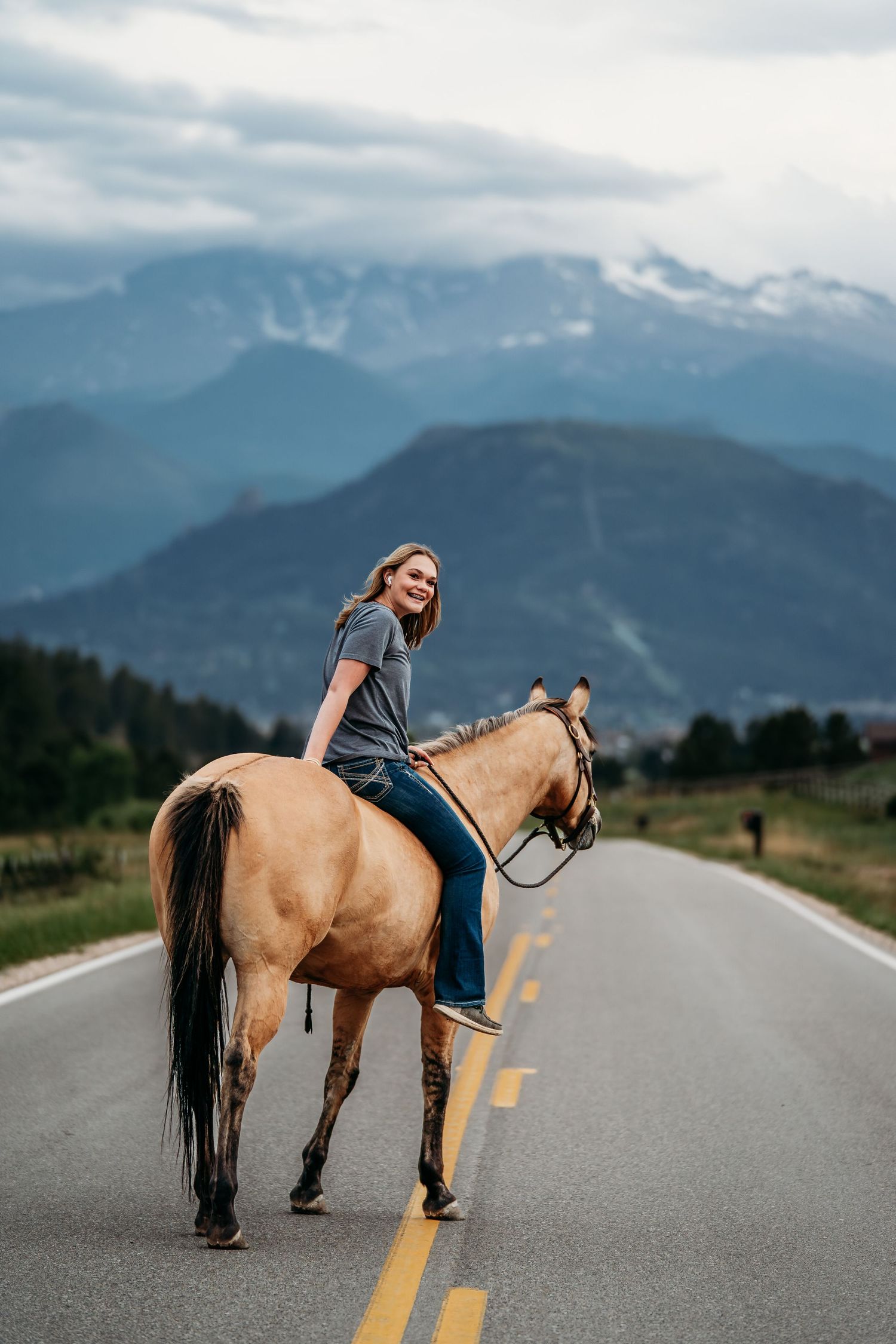 Woman riding a buckskin horse on a mountain road with scenic snow-capped peaks in the background.