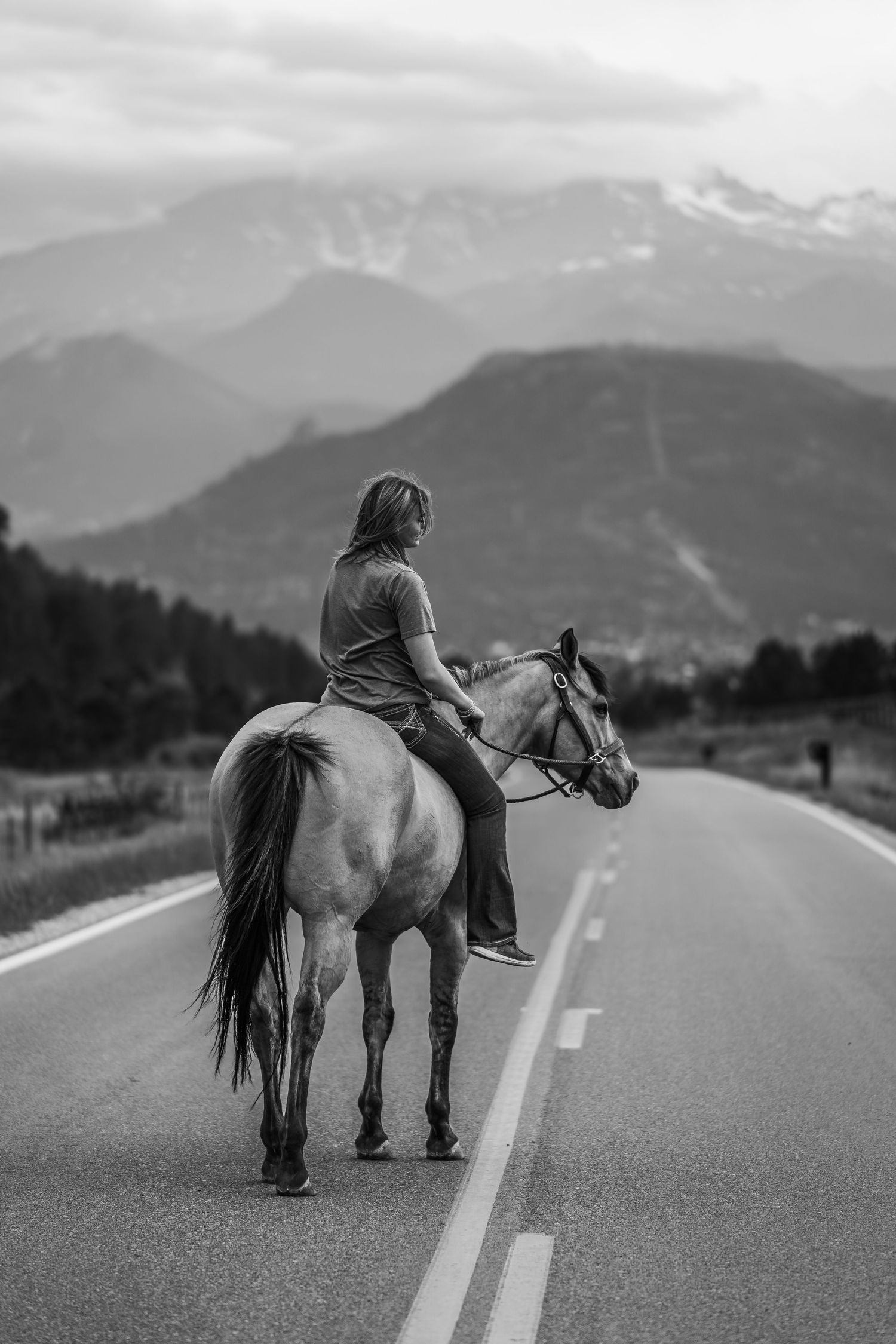 Black and white photo of a woman riding a horse on a mountain road with scenic peaks in the background.