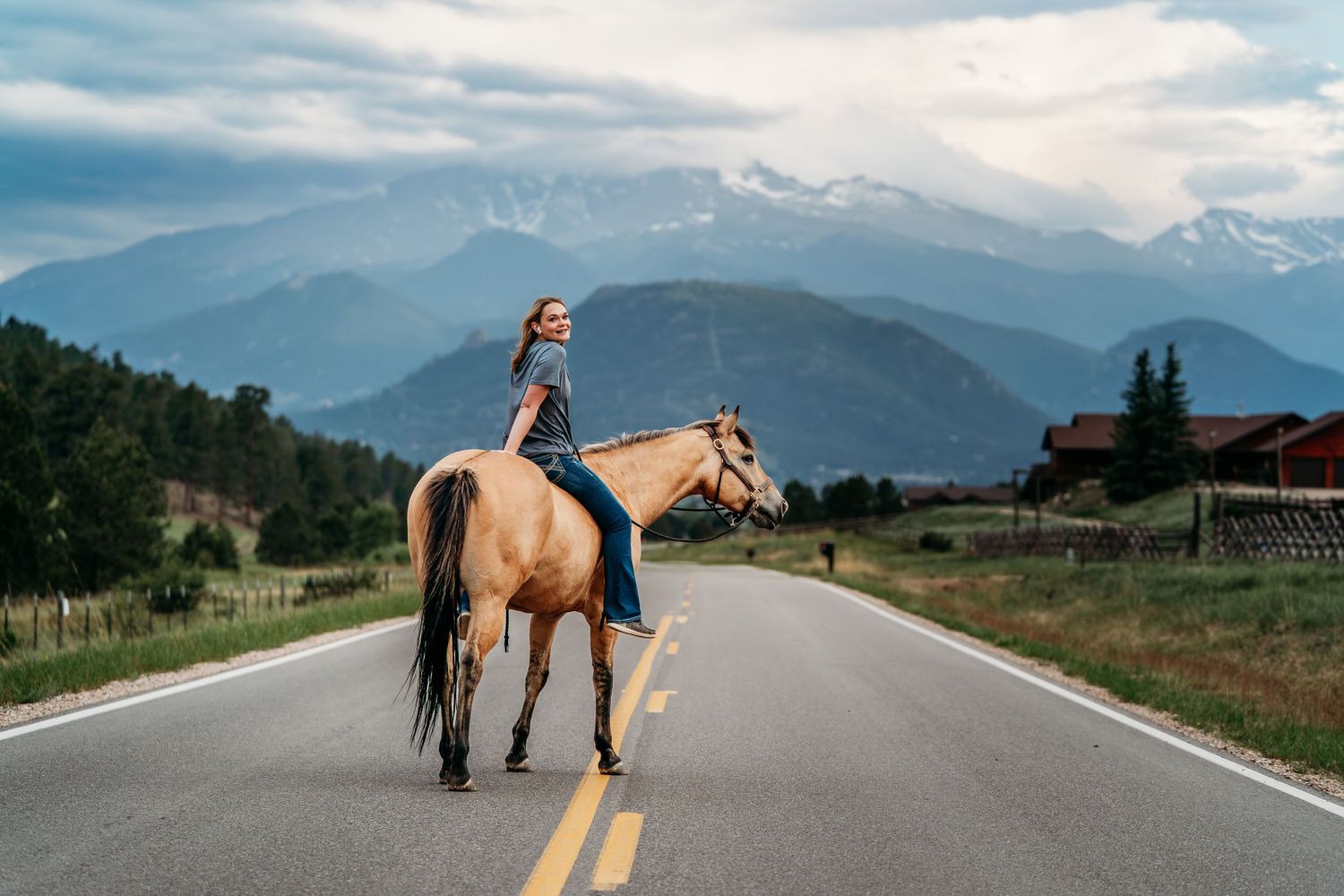 Woman riding a palomino horse on a mountain road with snow-capped peaks and dramatic cloudy skies in the background.