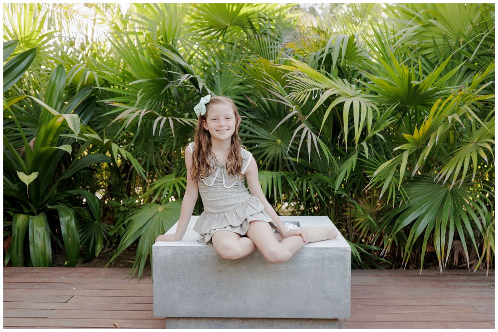 Young girl in gray dress sitting cross-legged on concrete bench surrounded by lush tropical palm trees.