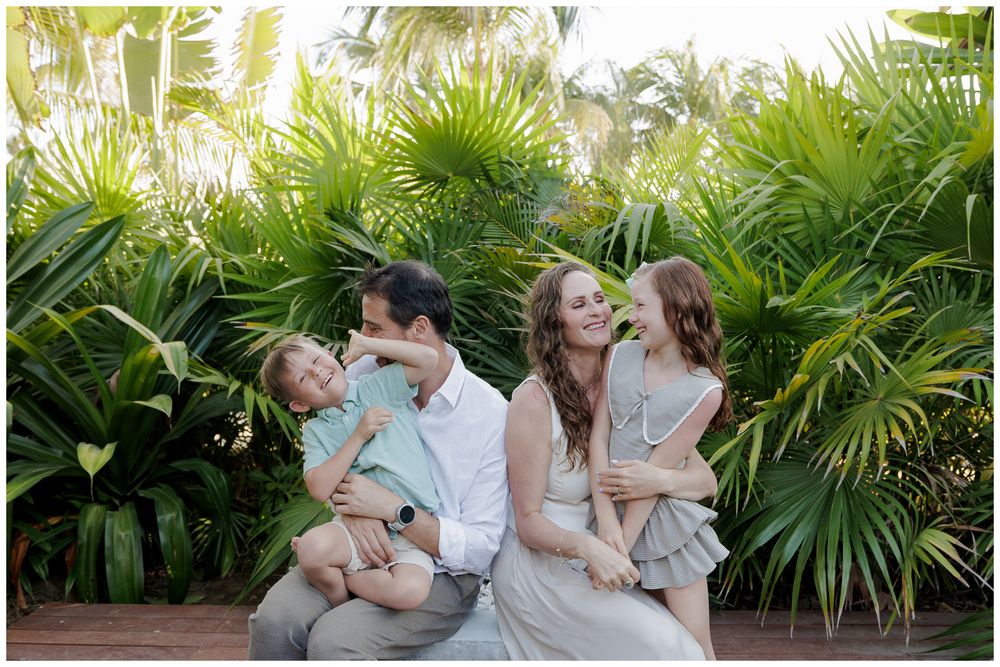 Happy family of four shares candid moment seated together in front of lush tropical greenery.