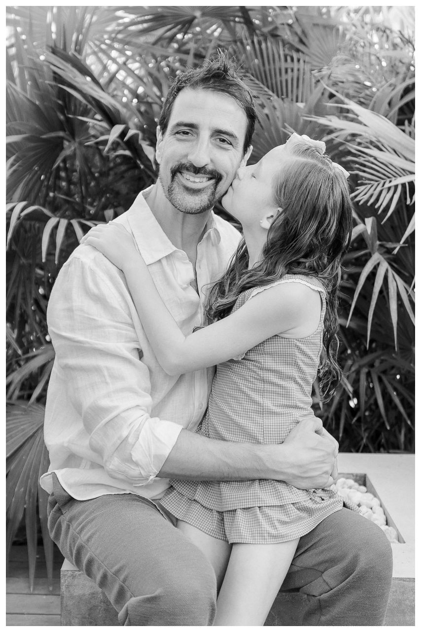 A smiling man with a mustache receives a sweet kiss on the cheek from a young girl outdoors.