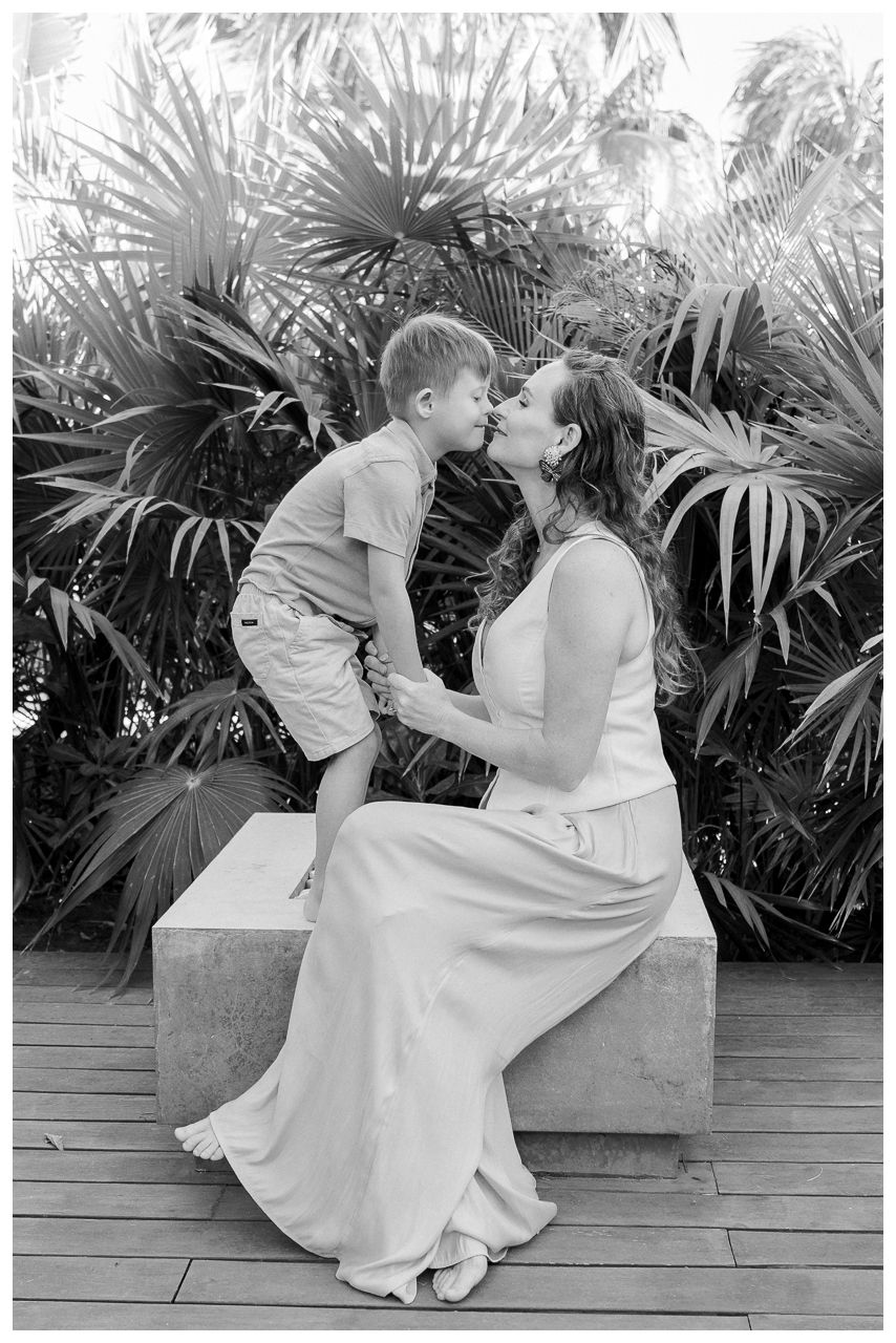 A young boy shares a tender kiss with a woman in a white dress seated outdoors amid lush palms.